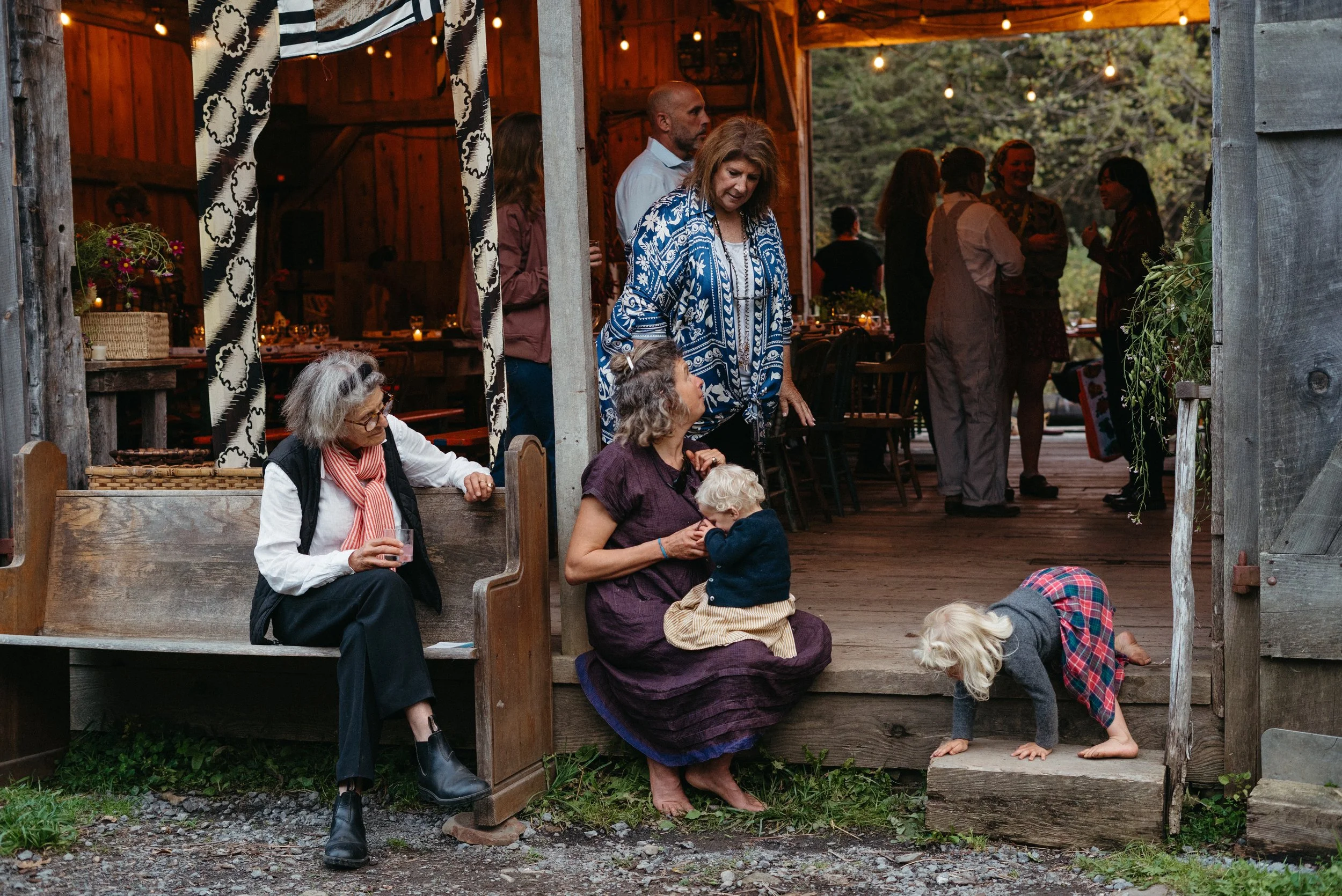 Group of people gathered on a wooden porch during an outdoor event, with some seated and others standing, including a woman with a child sitting on her lap and a small girl crawling on the ground.