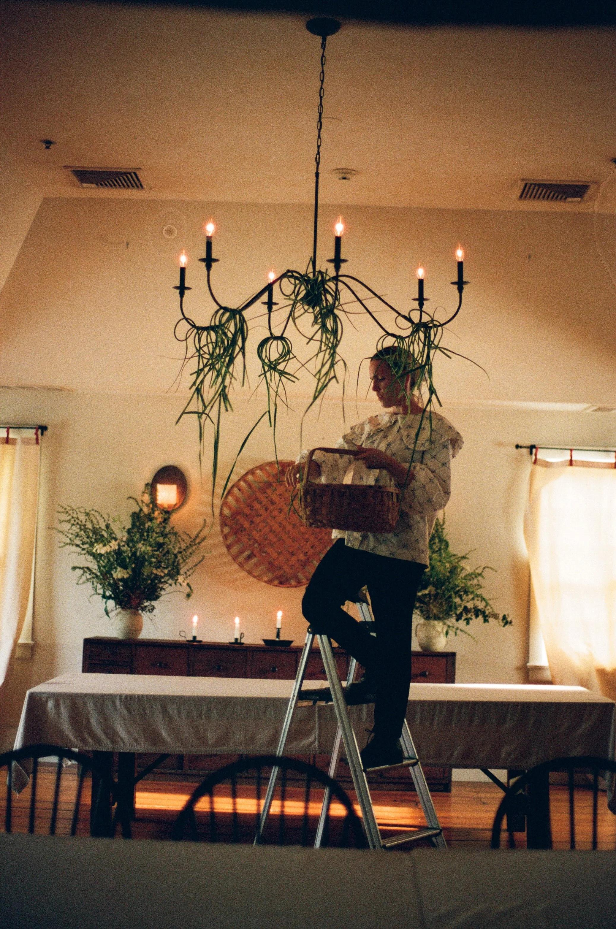 A woman standing on a ladder decorating a chandelier with plant branches in a dining room, with table, candles, and plants in the background.
