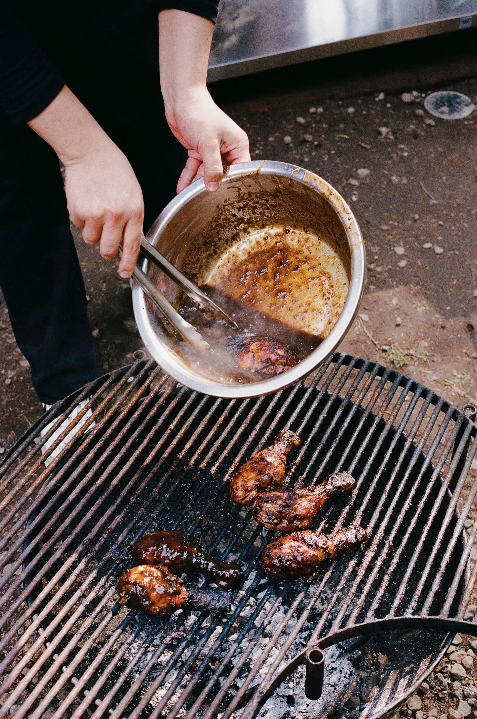 Person grilling chicken wings on a barbecue with a sauce brush, pouring sauce from a bowl onto the chicken.