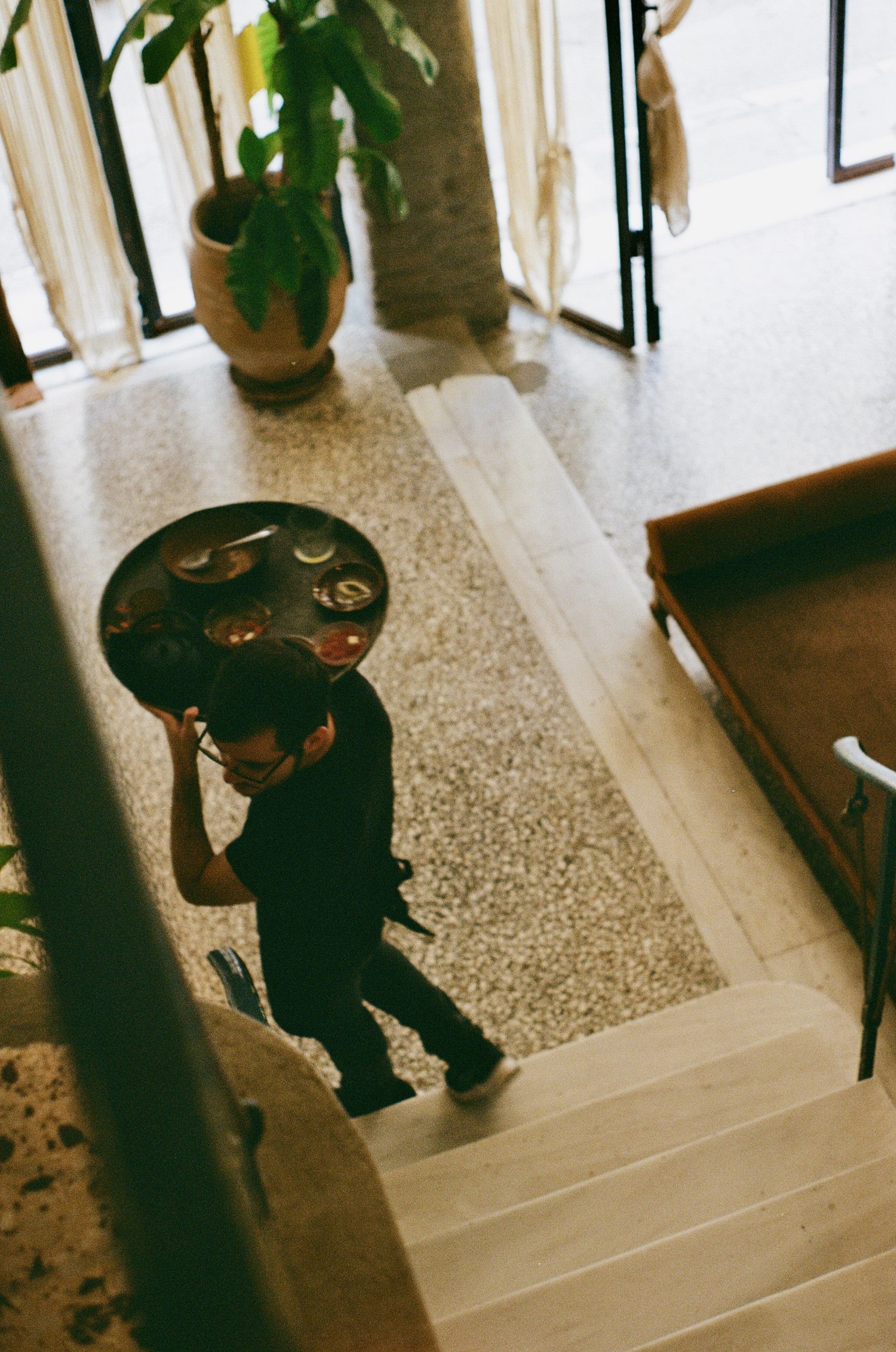 A person wearing glasses and a black shirt is carrying a tray with cups or glasses, walking down a staircase in a residential setting.