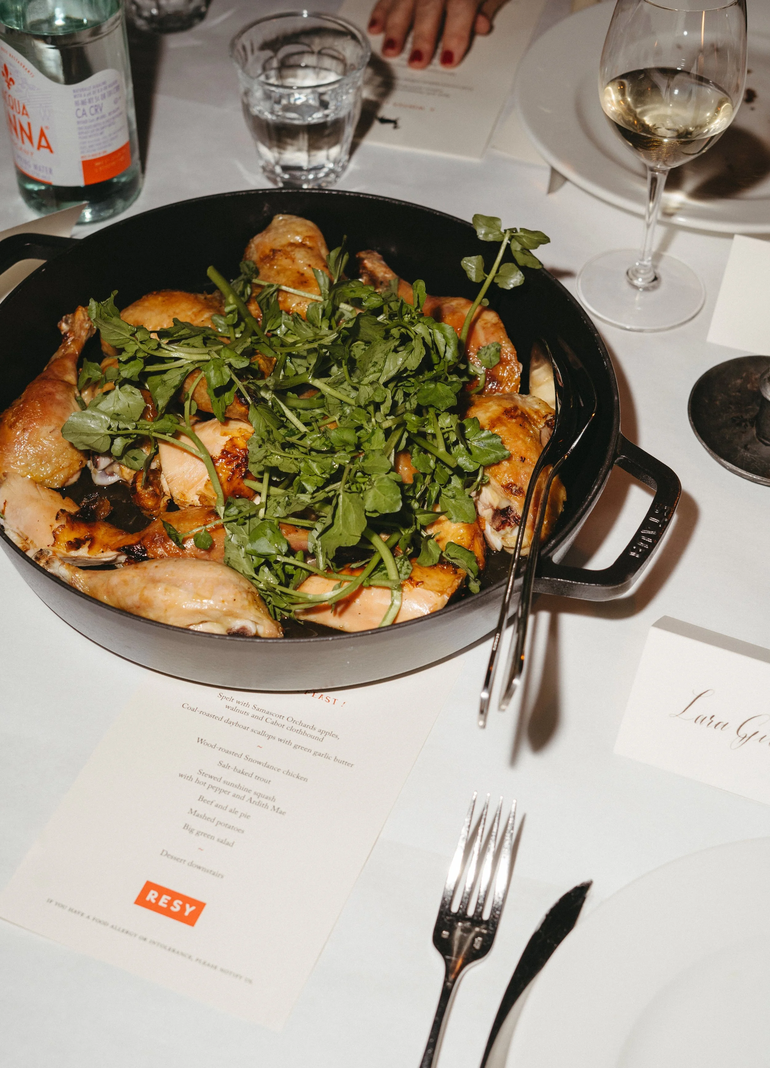 A table with roasted chicken garnished with fresh herbs, served in a black skillet, accompanied by a glass of white wine, water, a bottle of water, a fork and knife, and a printed menu on a white tablecloth.