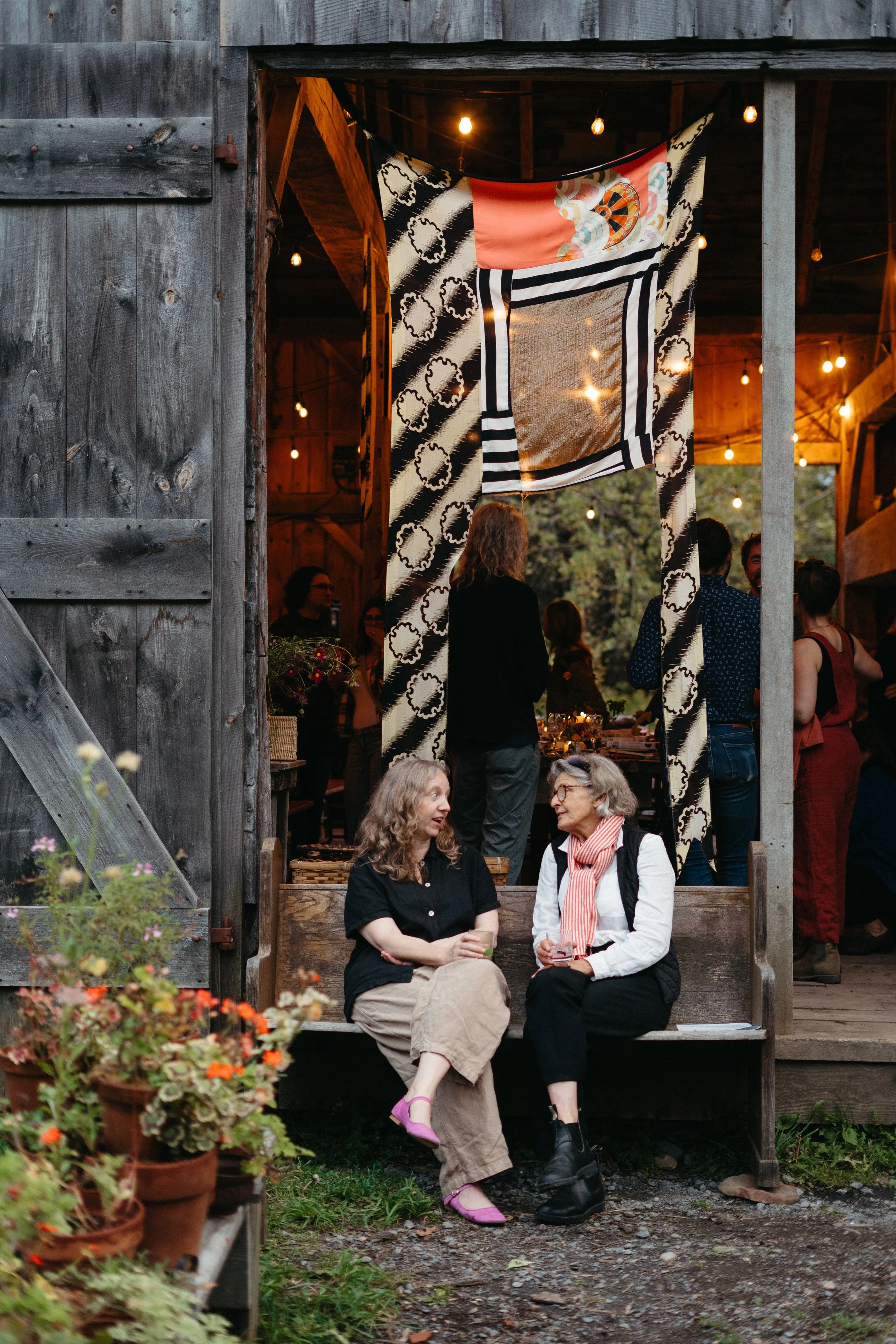 Two women are sitting and talking on a wooden bench outside a rustic barn, with a gathering of people inside, illuminated by warm string lights.