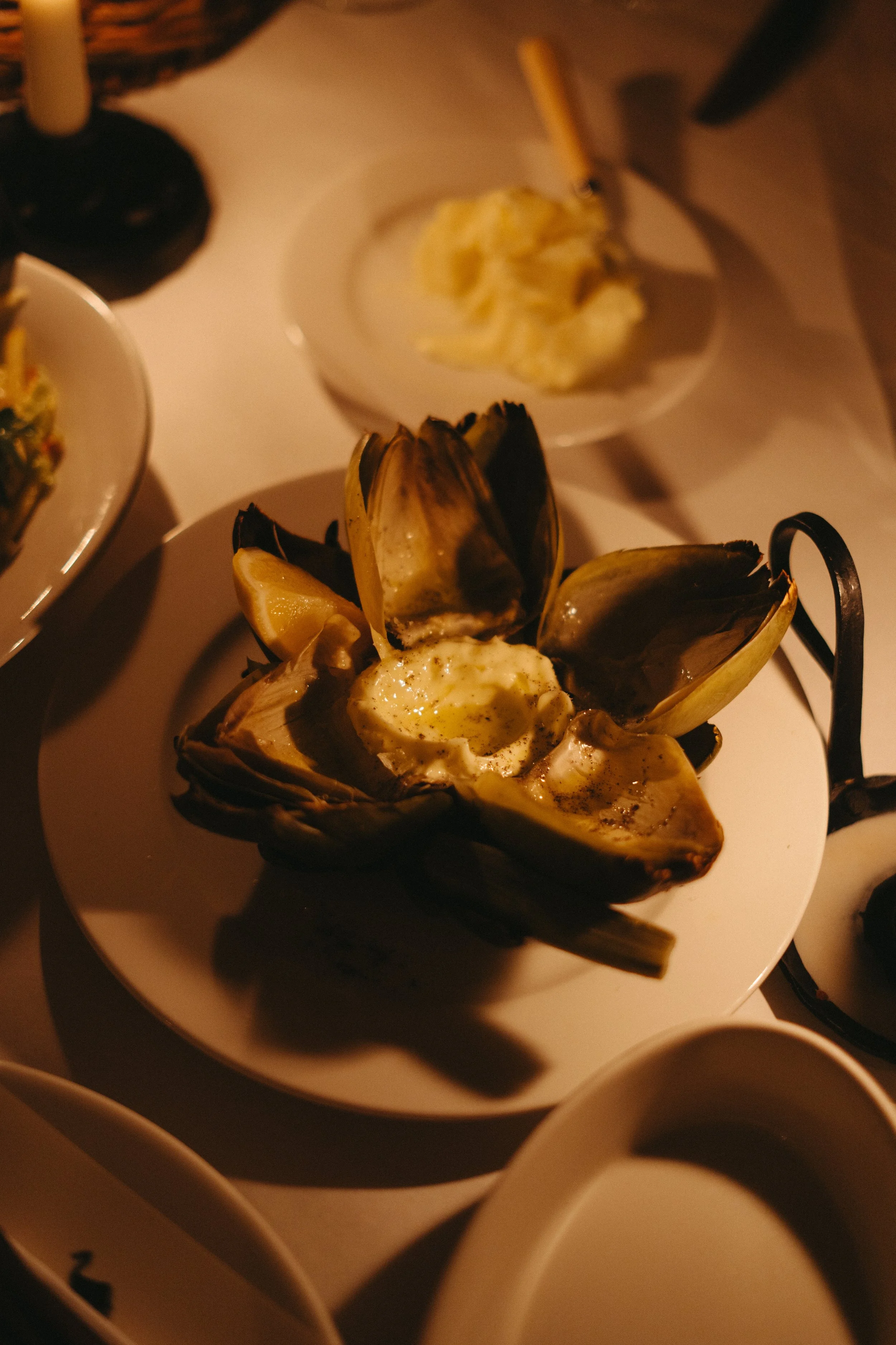A plate of cooked artichoke with melted butter or sauce, surrounded by other plates with food on a white tablecloth, dimly lit.