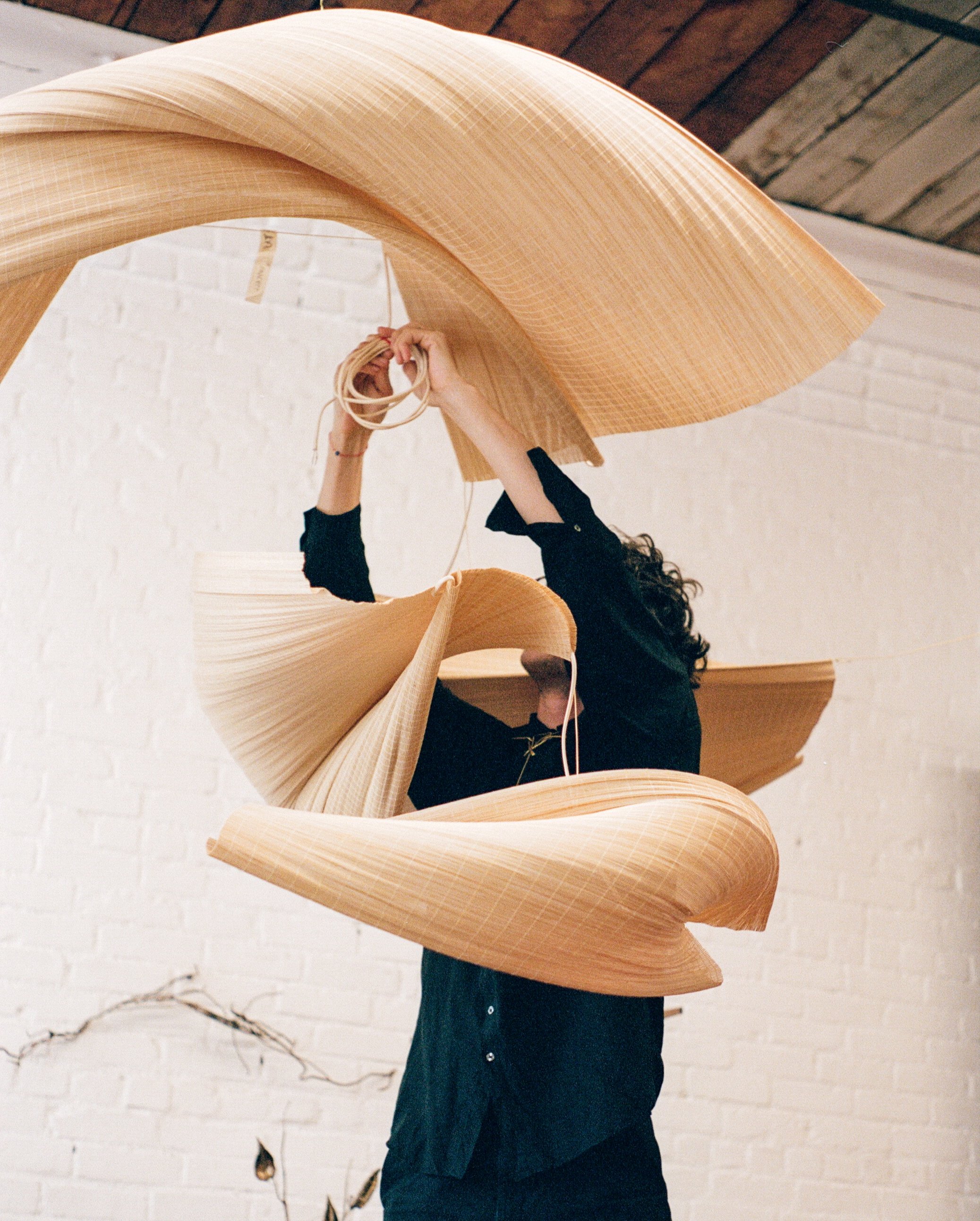 Person assembling or adjusting a large, abstract wooden sculpture or art installation with curved, layered wooden pieces in a room with white brick walls and a wooden ceiling.