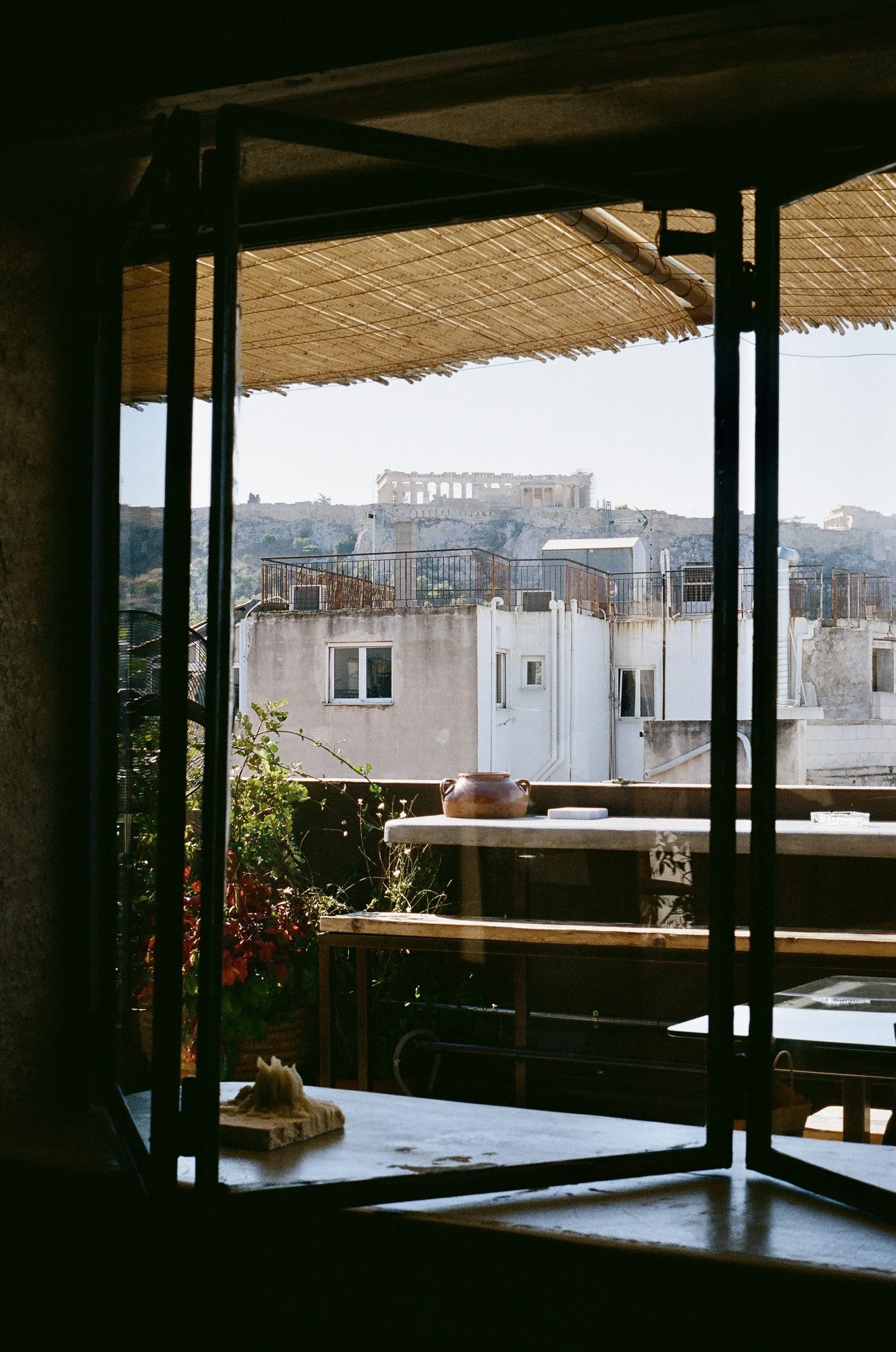 View of the Acropolis in Athens, Greece, through a window with a bamboo shade overhead, showing white buildings and a clear sky.