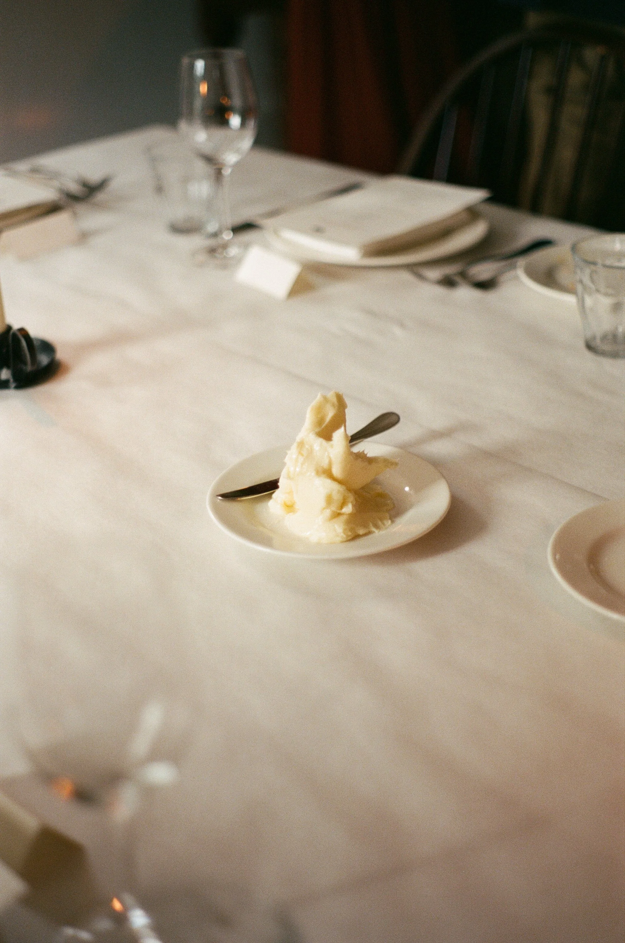 A small white plate with a scoop of vanilla ice cream and a spoon on a dining table set for a meal.