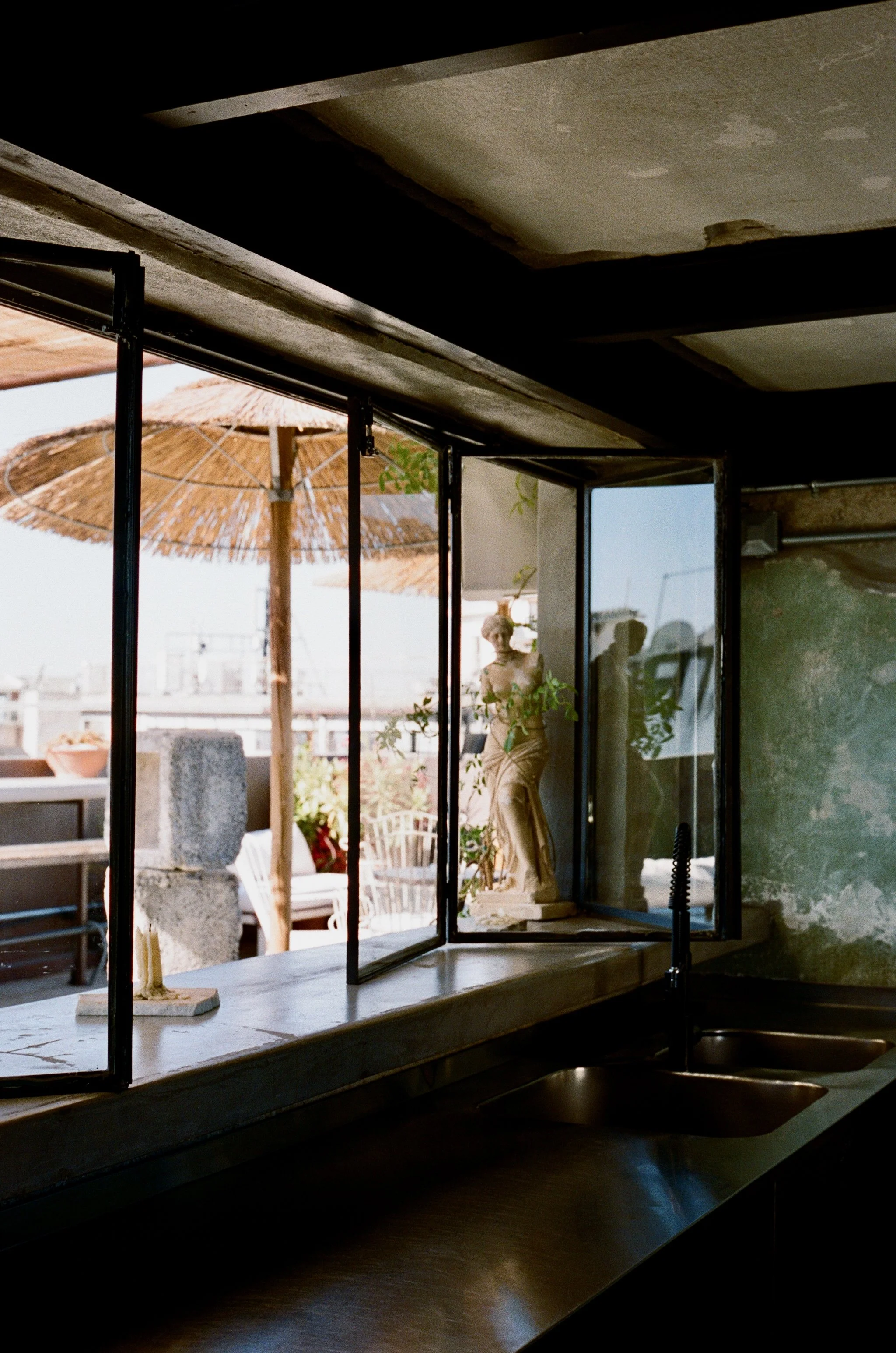 View from inside a kitchen looking out a large open window with black metal frame, revealing a patio with a thatched umbrella, potted plants, and a classical marble statue on a pedestal.