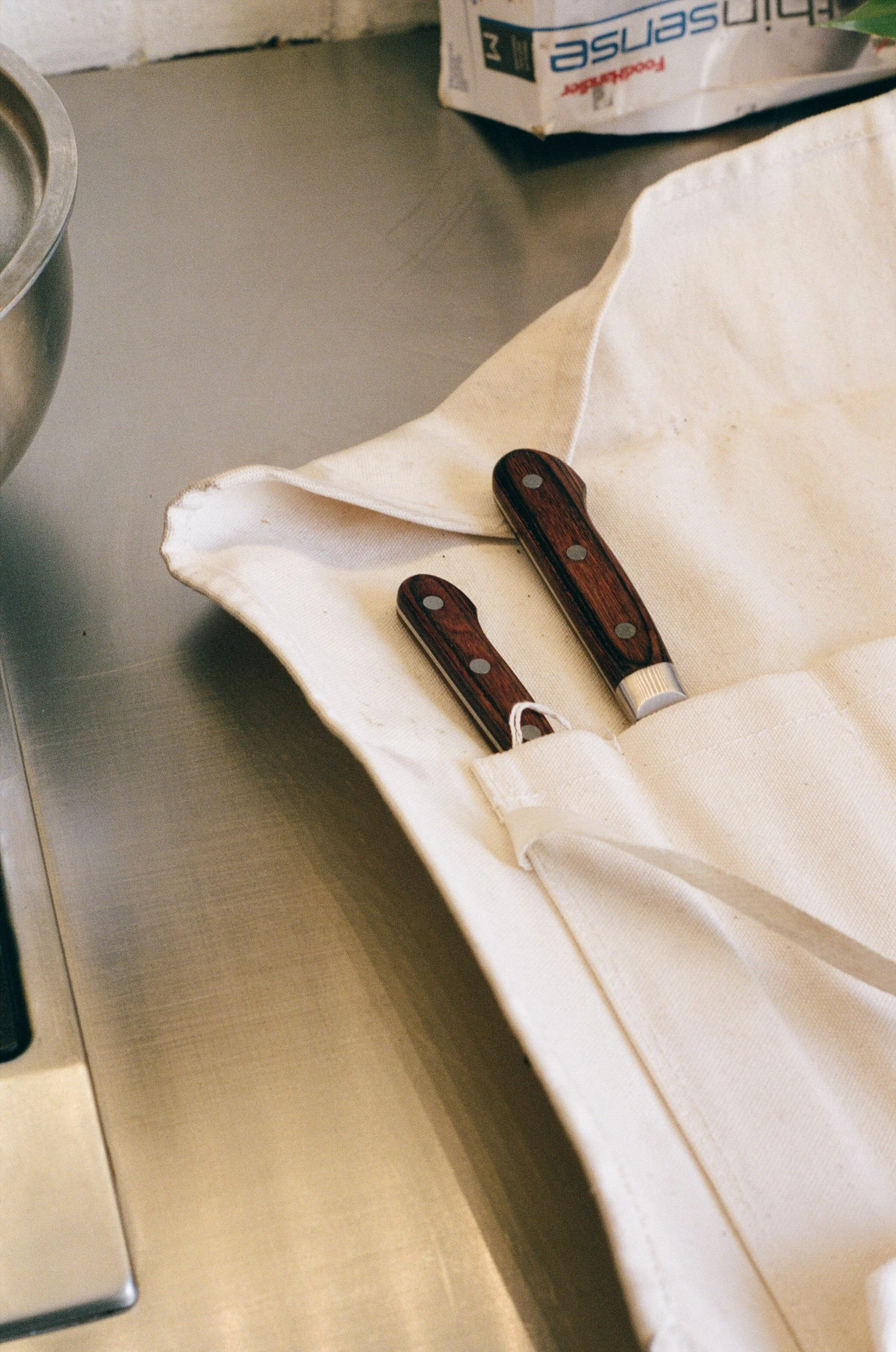 Two kitchen knives with wooden handles resting in a white cloth on a stainless steel countertop.