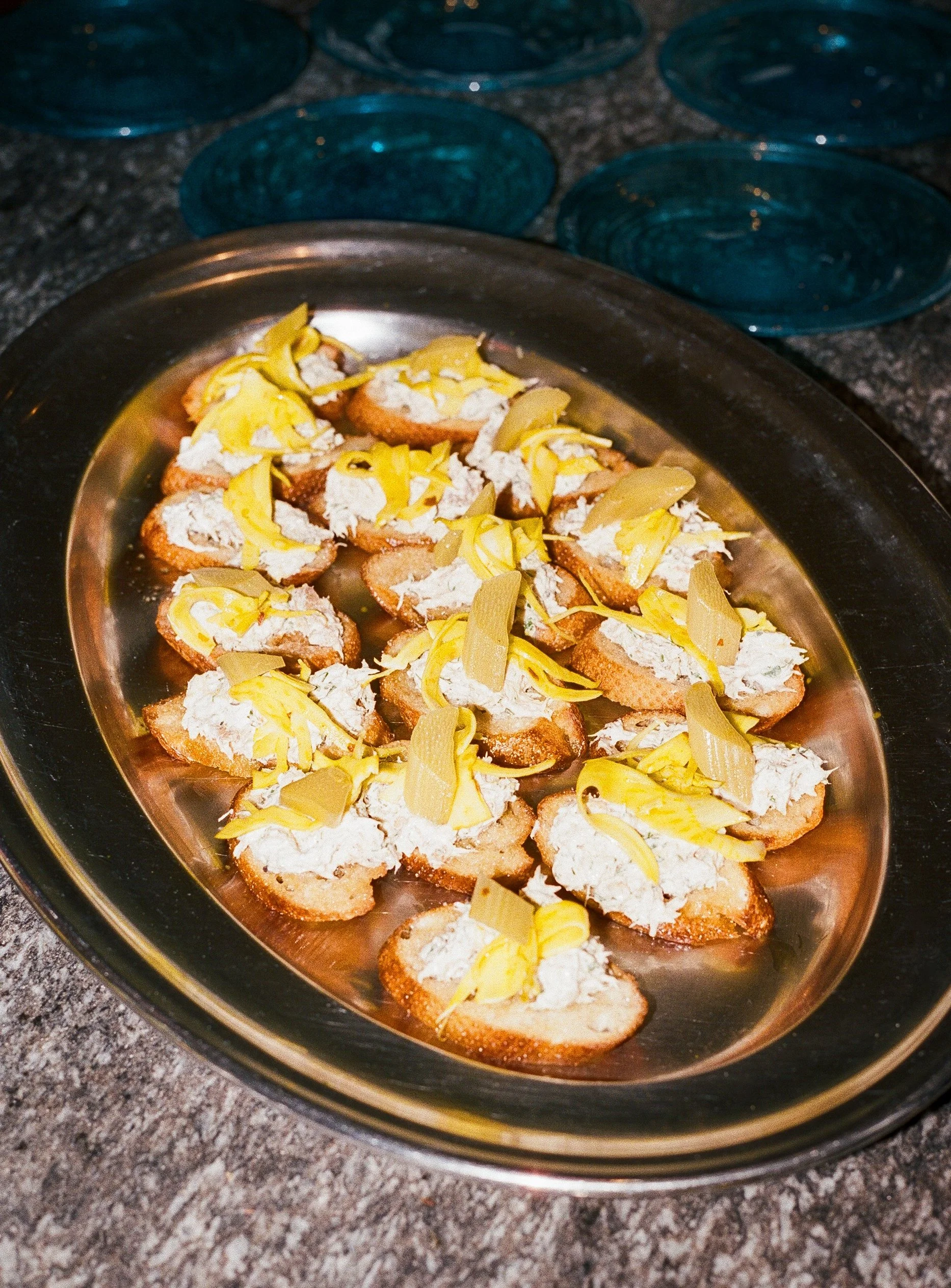 Plate of small bread slices topped with white cheese and yellow pickles, set on a silver platter on a dark countertop with blue glass bowls in the background.