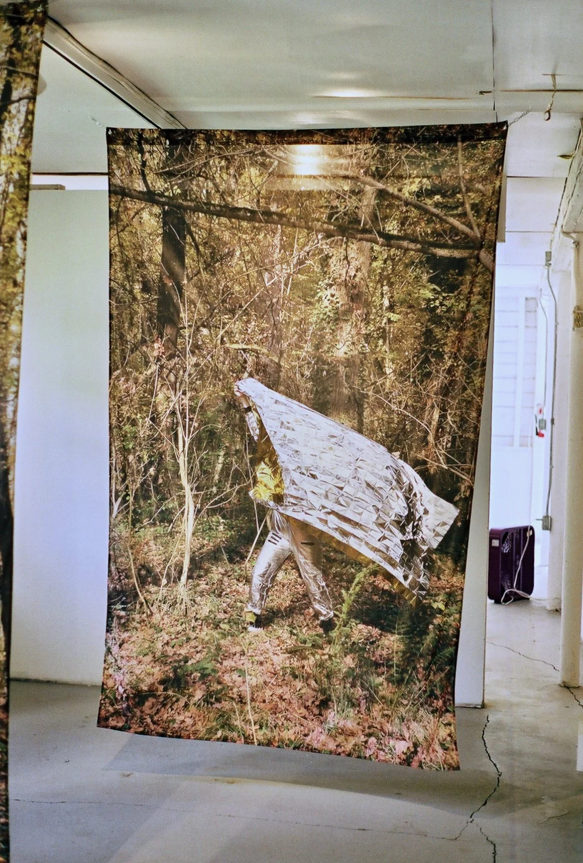 An art installation featuring a large printed fabric backdrop of a forest, with a person partly covered in shiny metallic material walking through the scene.