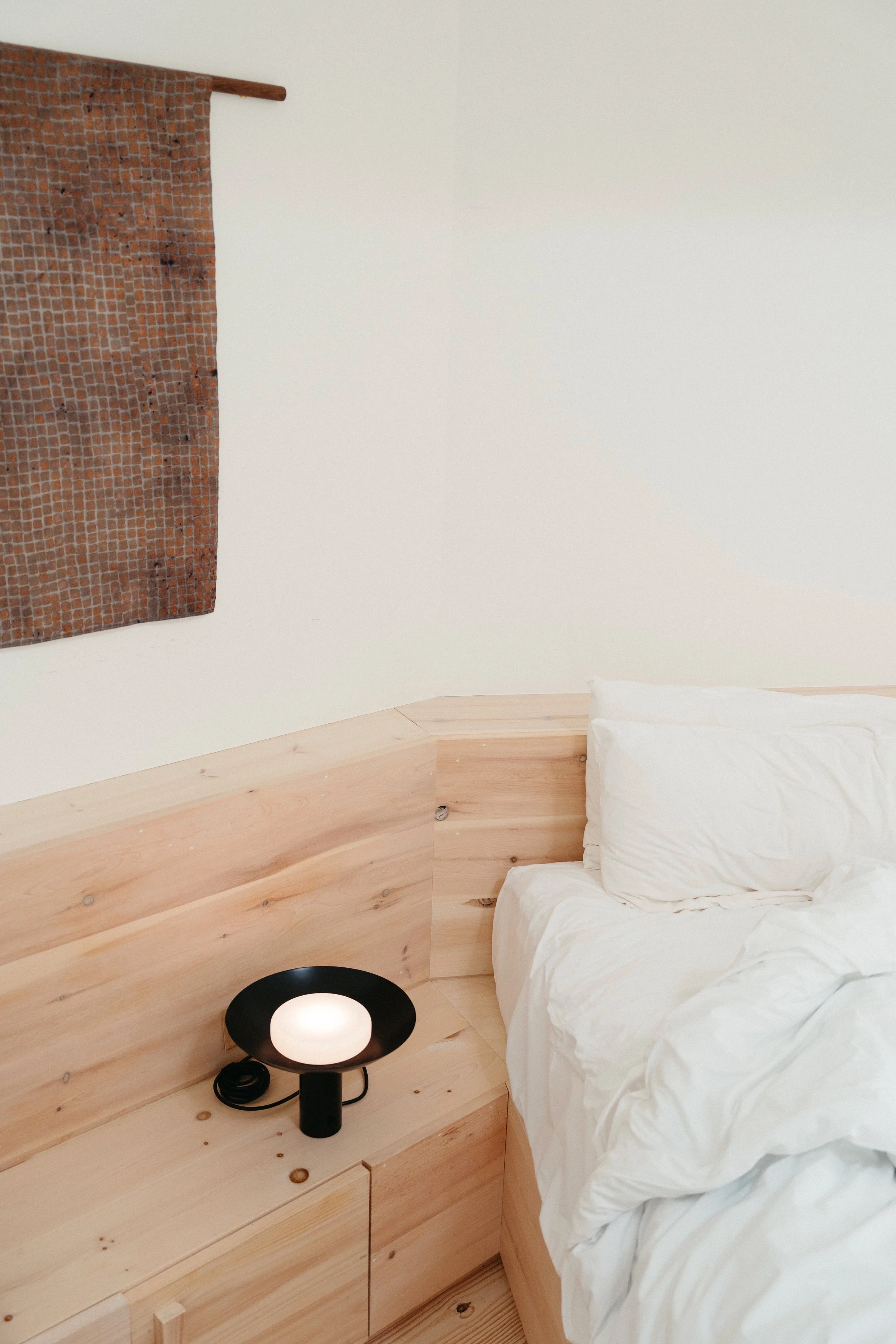 A hotel room with a bed, white bedding, and a wooden headboard. There is a modern black lamp on a wooden nightstand, and a decorative brown woven wall hanging on the white wall.