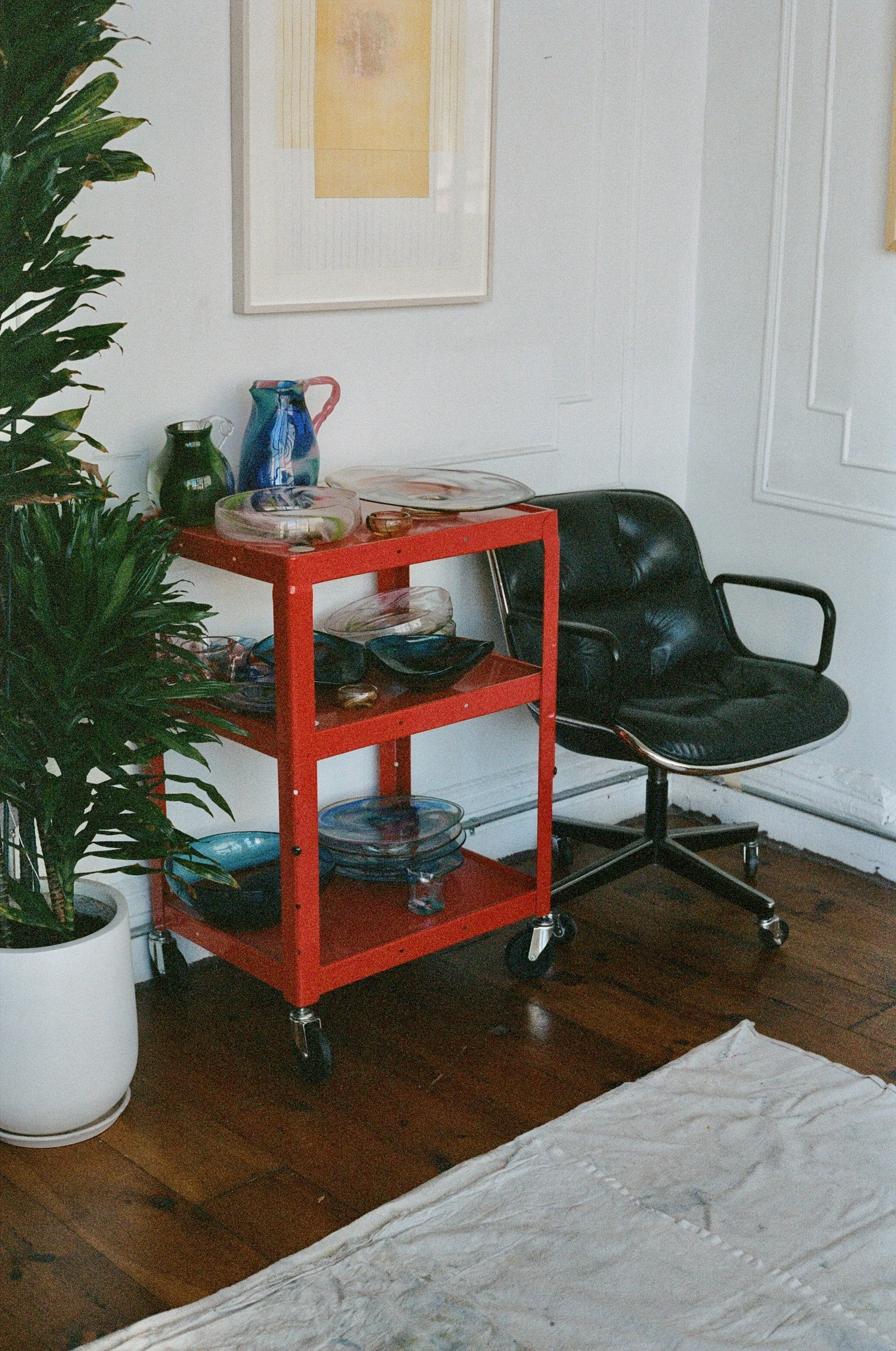 A corner of a room with a white wall, a black leather office chair, a red rolling cart with glass serving dishes, a green potted plant in a white pot, and framed artwork hanging on the wall.