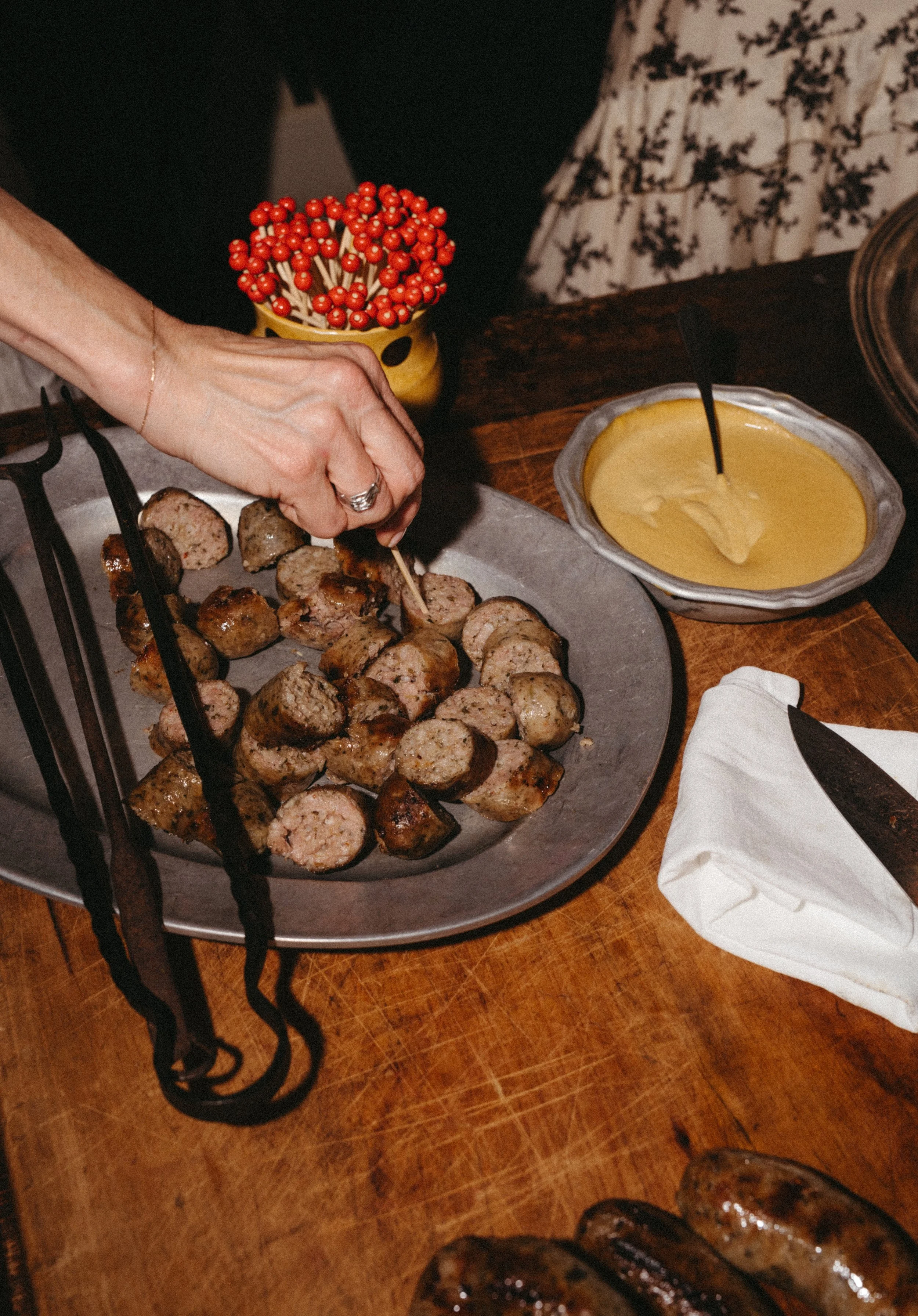 Person cutting cooked sausage on a large silver platter; bowl of mustard and a rolled cloth napkin with a knife on it on a wooden table; small bowl of red berries in background.