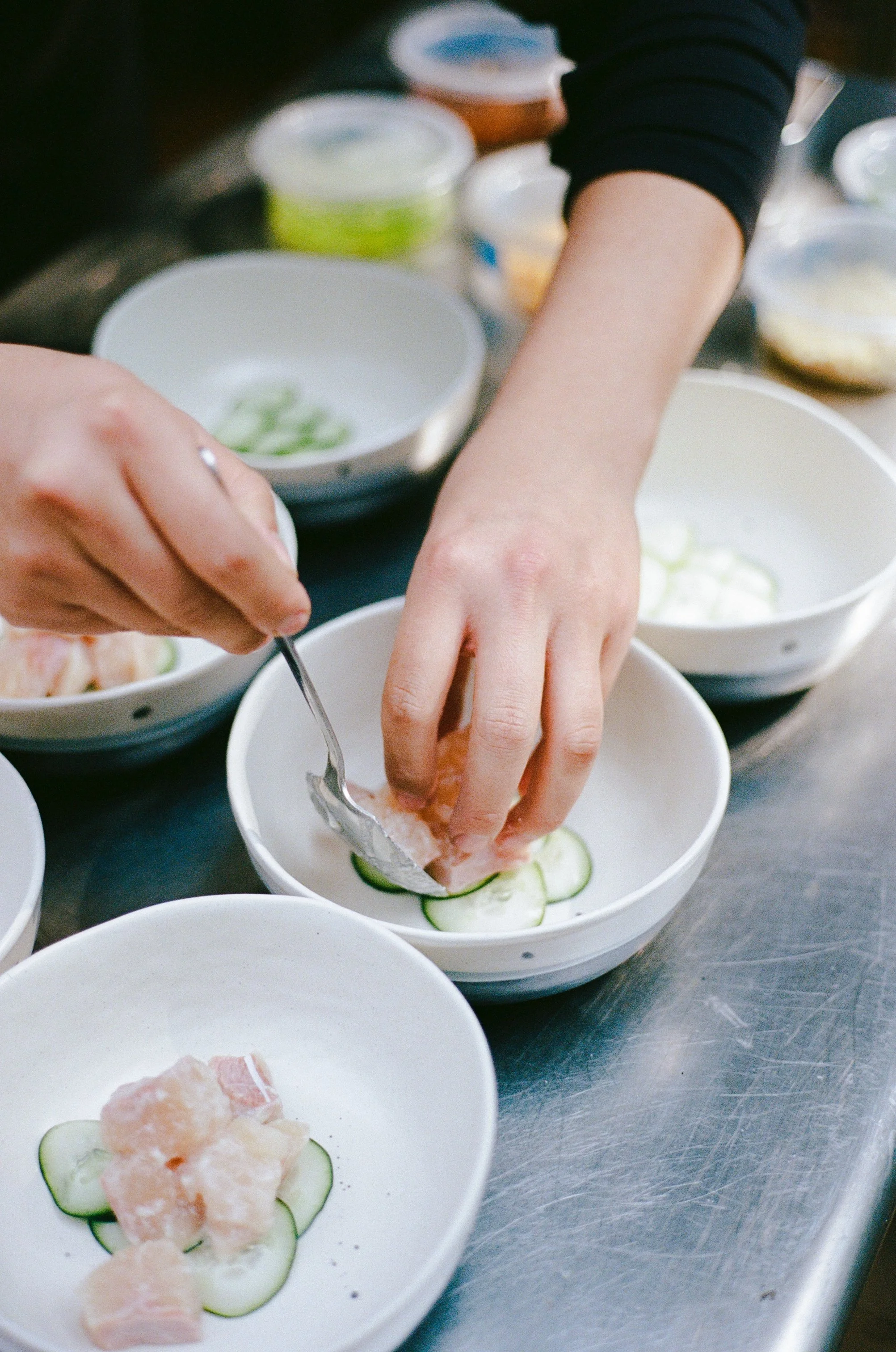 Person assembling sushi, placing cucumber slices and raw fish into small white bowls, with various food containers in the background.