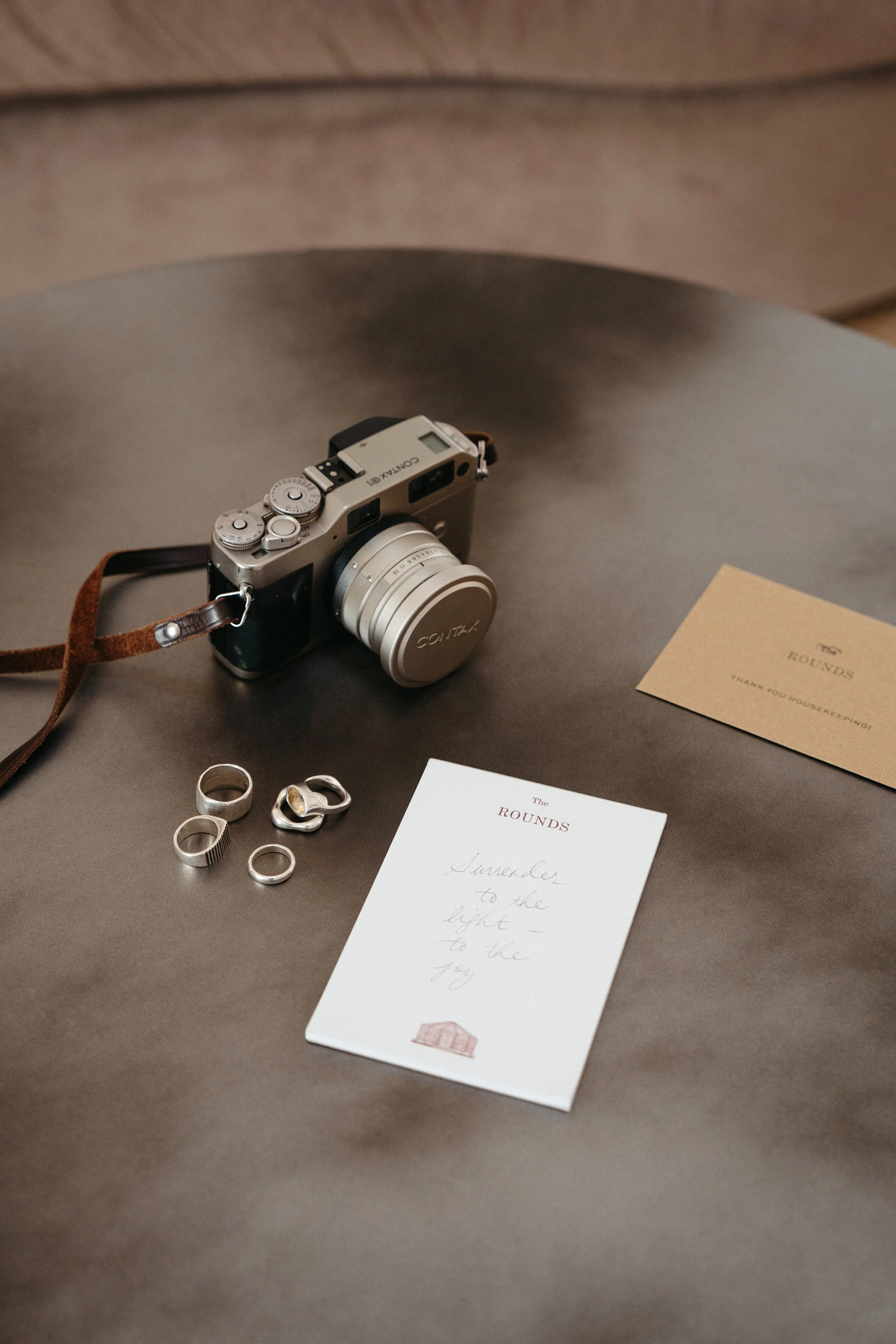 A vintage-style camera with a brown leather strap, four silver rings, a notepad titled 'The Rounds' with handwritten text, and a beige card on a dark surface.