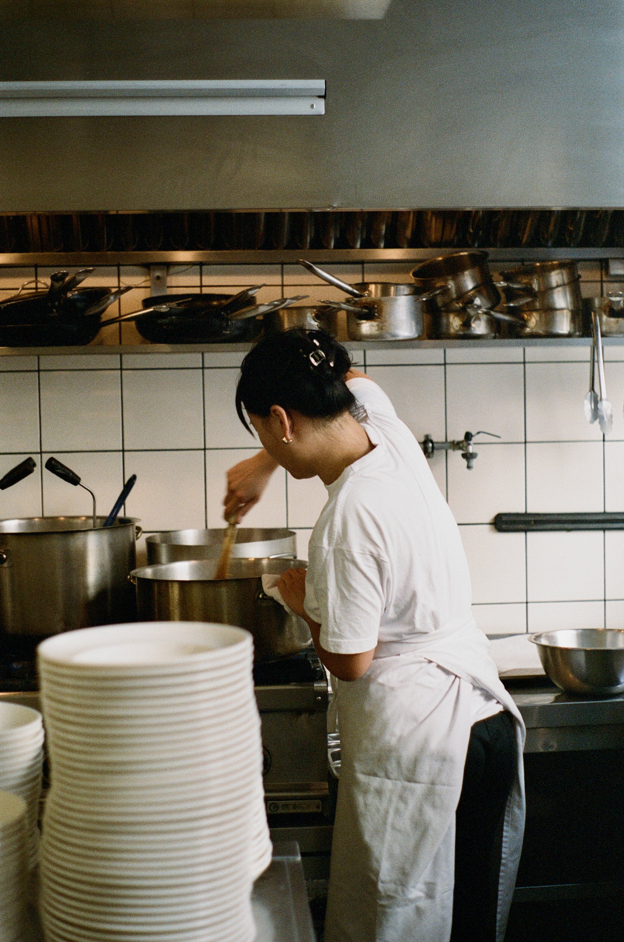 A person cooking in a commercial kitchen, stirring a pot on the stove surrounded by other cookware and dishes.