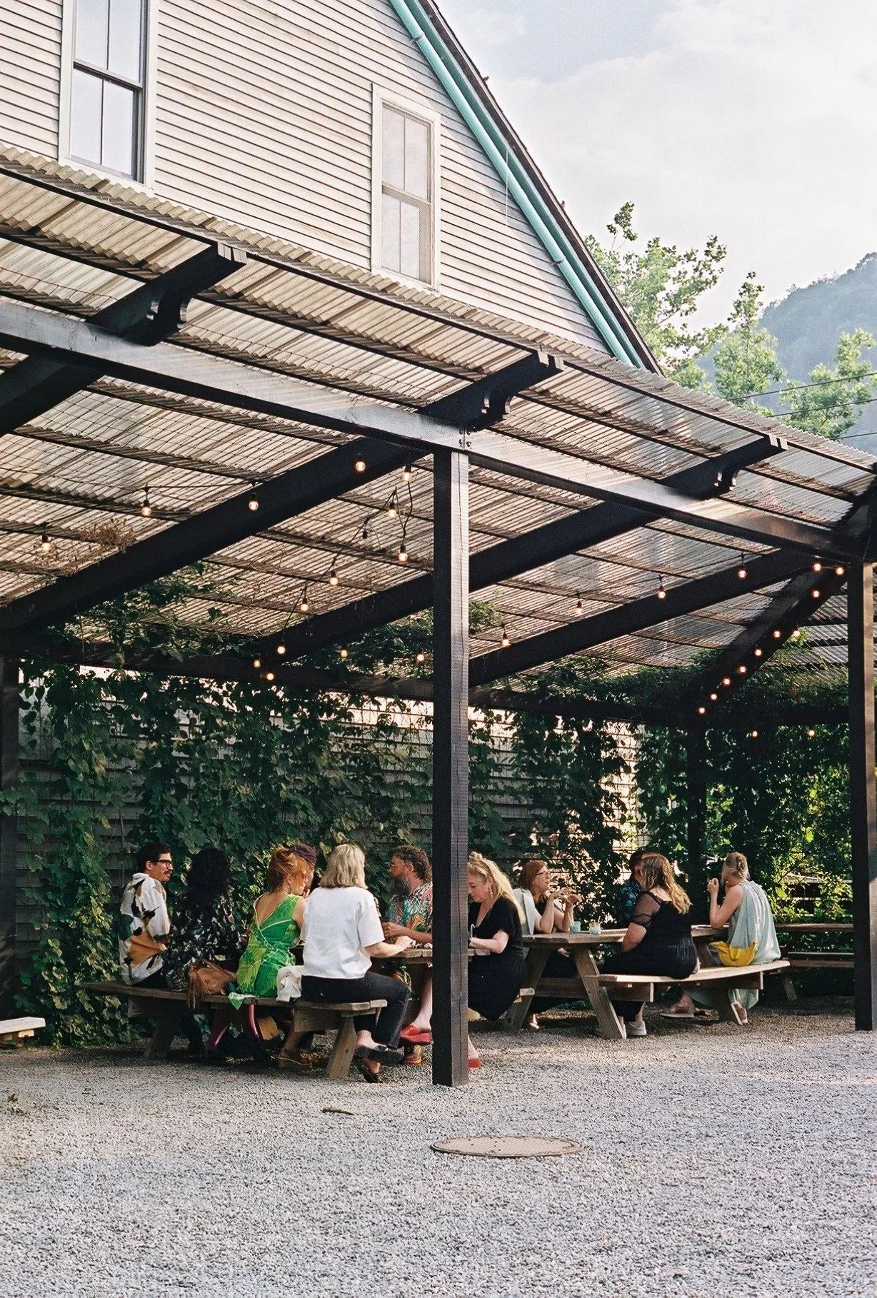 Group of people sitting at picnic tables under a pergola with string lights, adjacent to a beige house with green trim, with trees and mountains in the background.