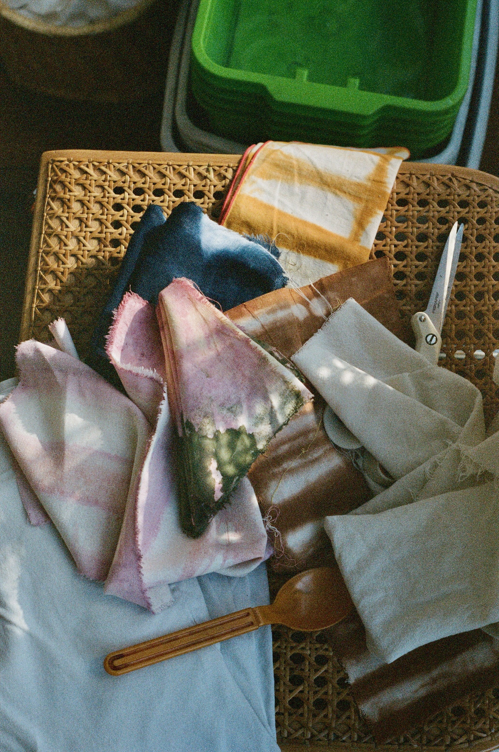 Assorted fabric pieces, a wooden spoon, and scissors on a woven surface with a green container in the background.