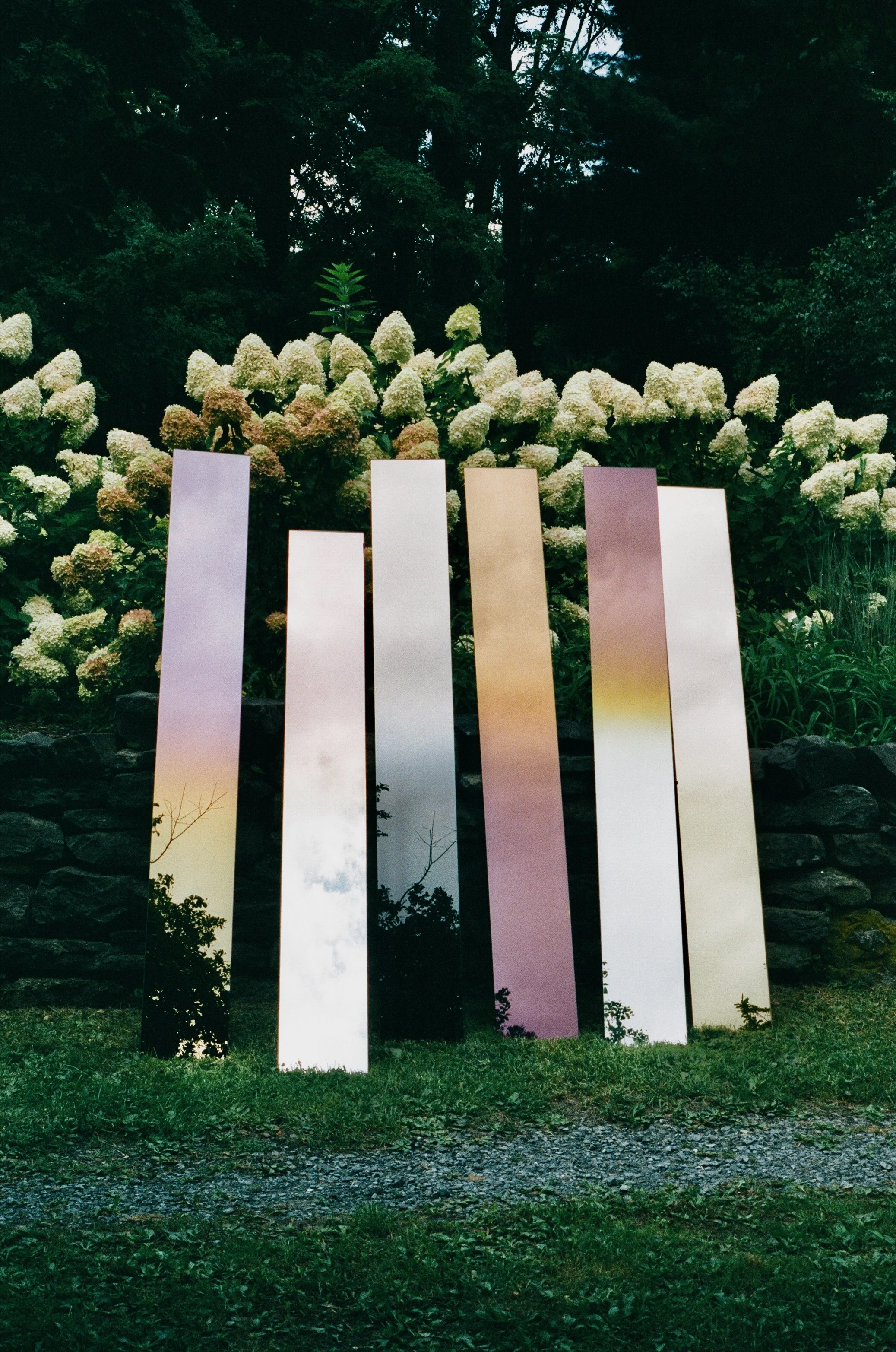 Reflective modern sculpture with vertical panels in front of a lush garden with white flowers and trees.