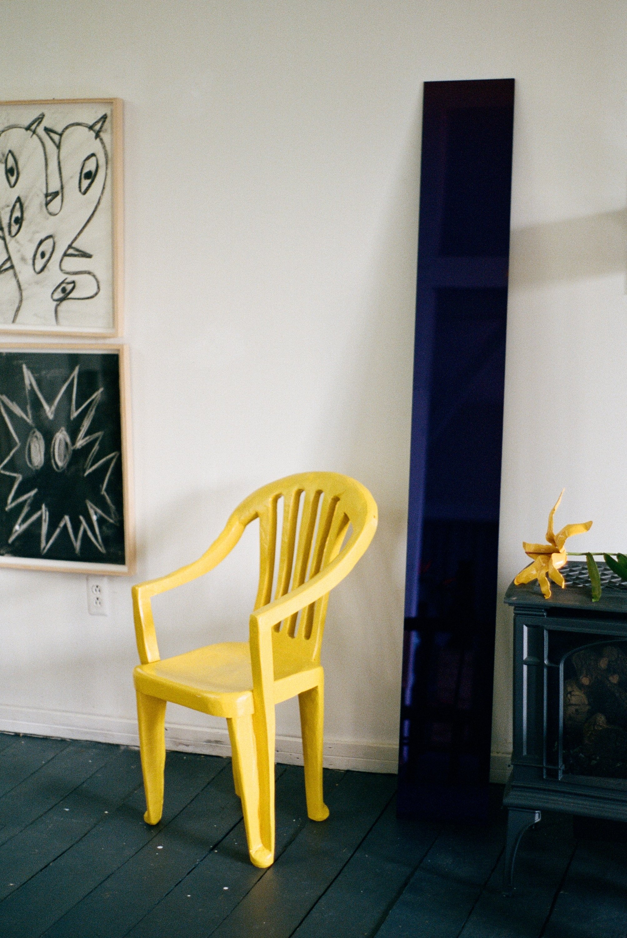 A yellow wooden chair next to a tall black mirror or panel in a room with white walls, dark wooden floors, and framed abstract art on the wall.