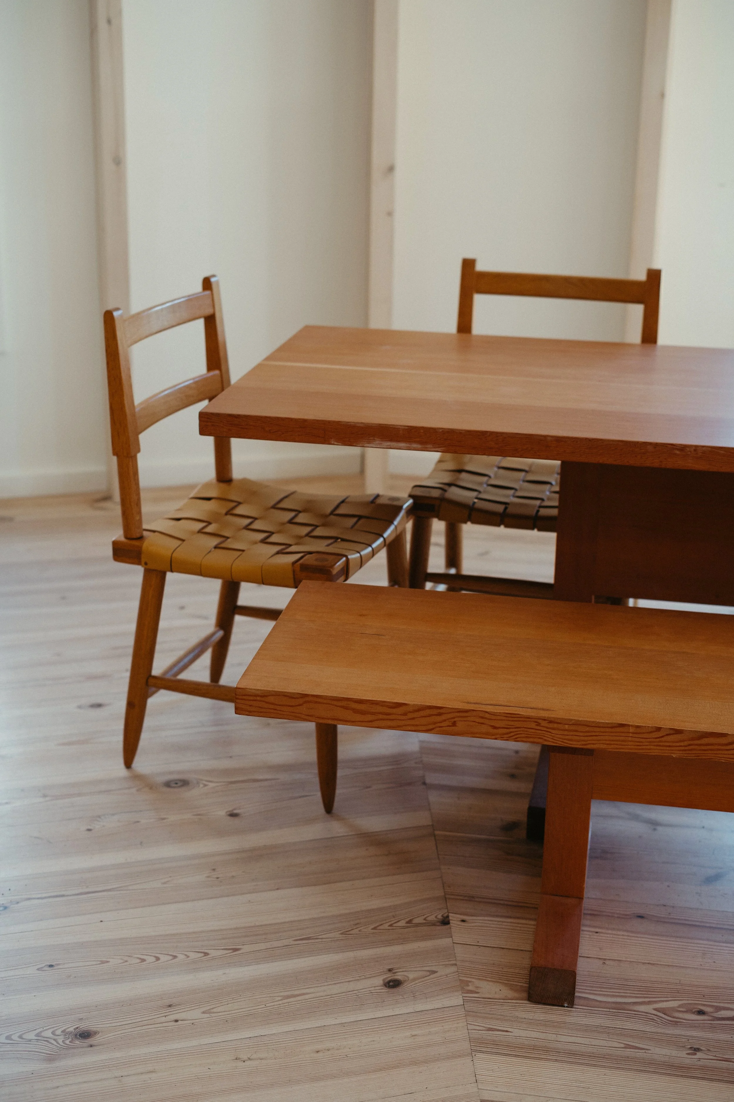 Interior of a room with wooden flooring and furniture, including a wooden table, two wooden chairs with woven seats, and a wooden bench.