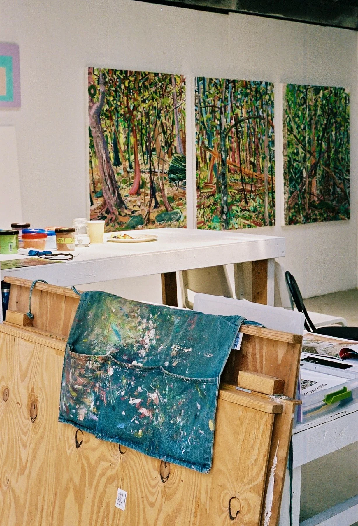 An art studio with three paintings of a forest scene hung on the wall, a wooden partition with a paint-splattered cloth, and art supplies on a white table.