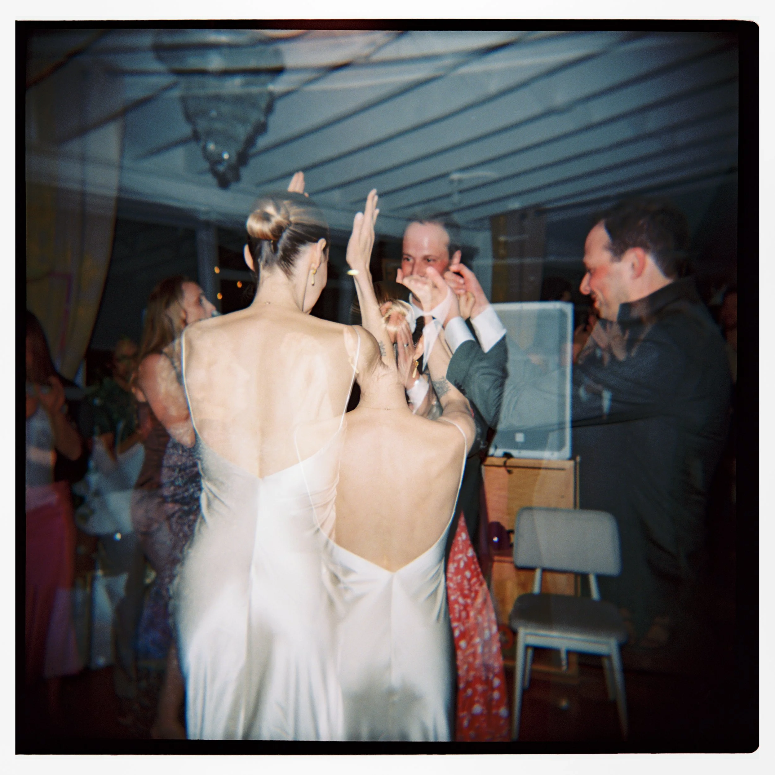People dancing and celebrating at a wedding reception, with women in elegant dresses and men in suits, inside a decorated venue.