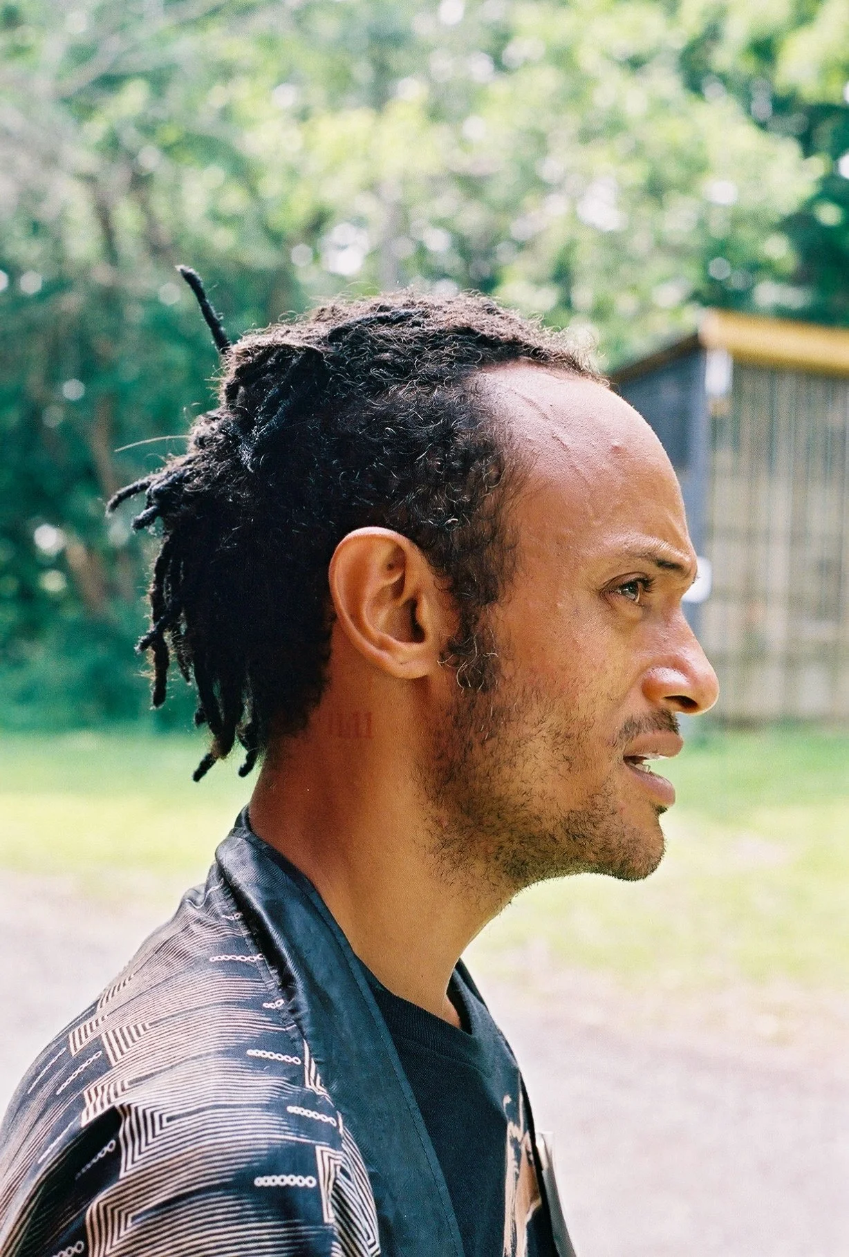 Profile of a man with dreadlocks, wearing a patterned jacket, outdoors with green trees in the background.