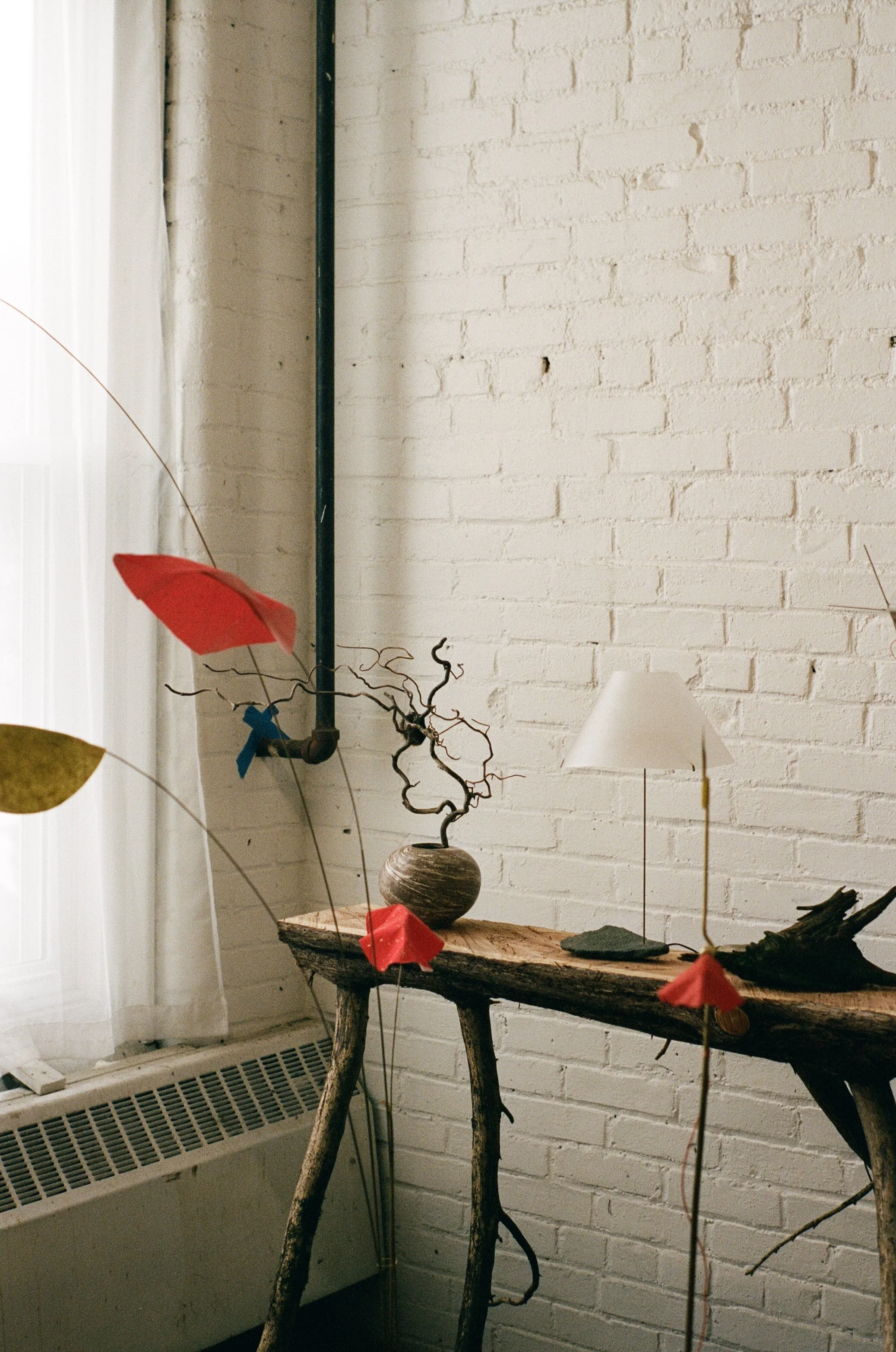 Decorative arrangement on a rustic wooden table against a white brick wall, including abstract twisted metal sculpture, a small white lamp, and colorful paper or fabric flowers on thin stems.