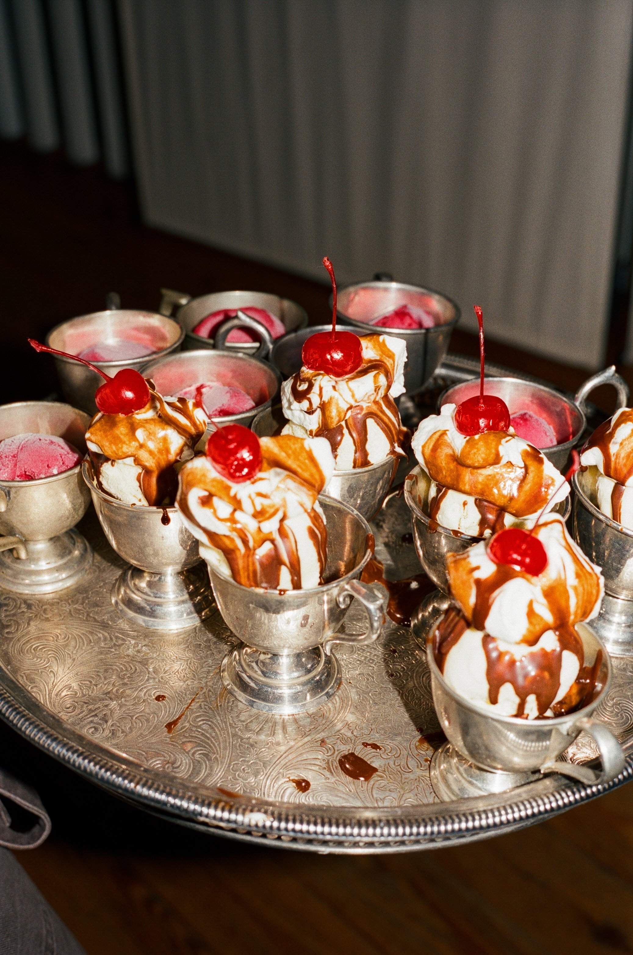 Ice cream sundaes topped with whipped cream, caramel sauce, and cherries served in small silver cups on a vintage tray.