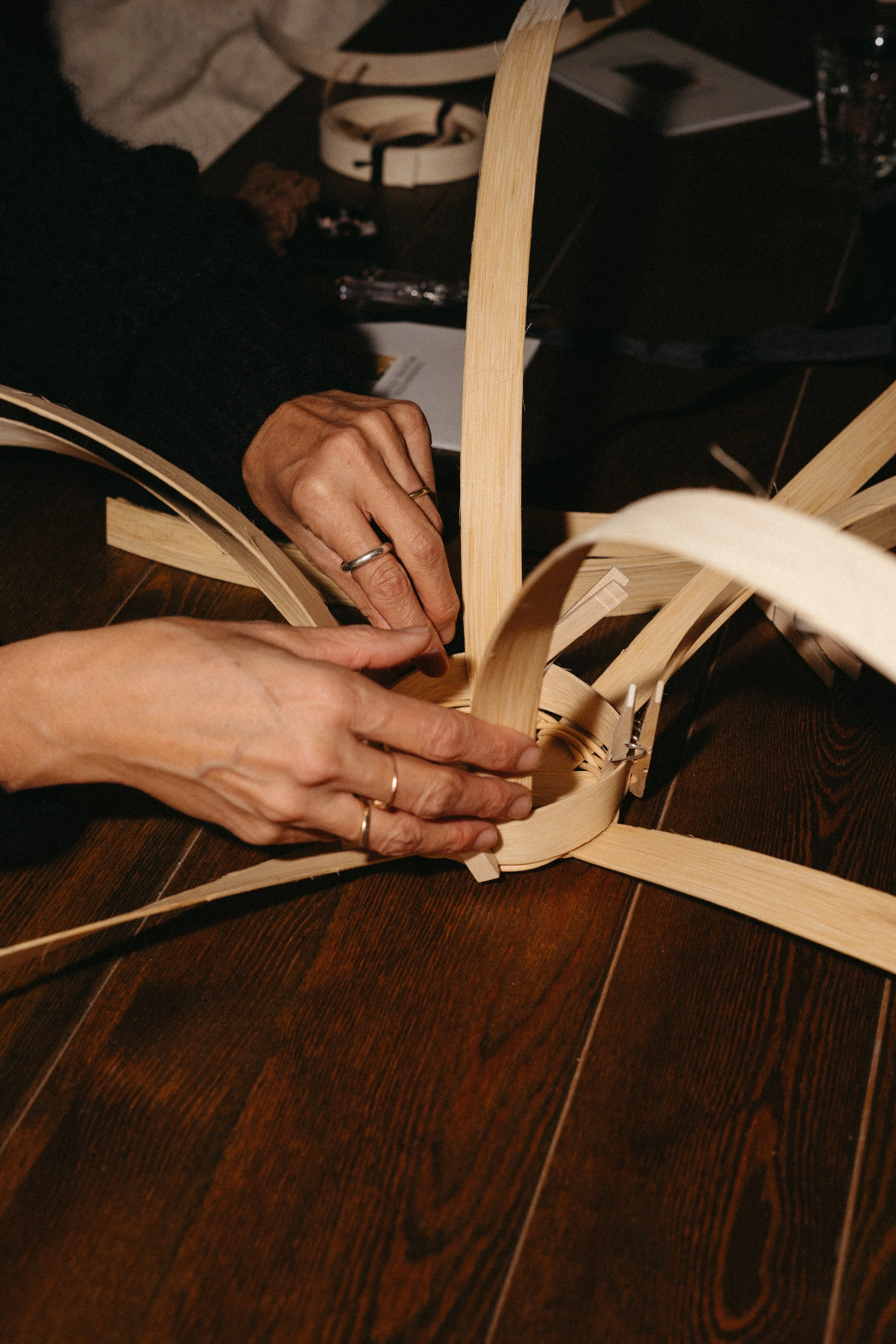 Hands assembling a woven wooden sculpture on a dark wooden table.