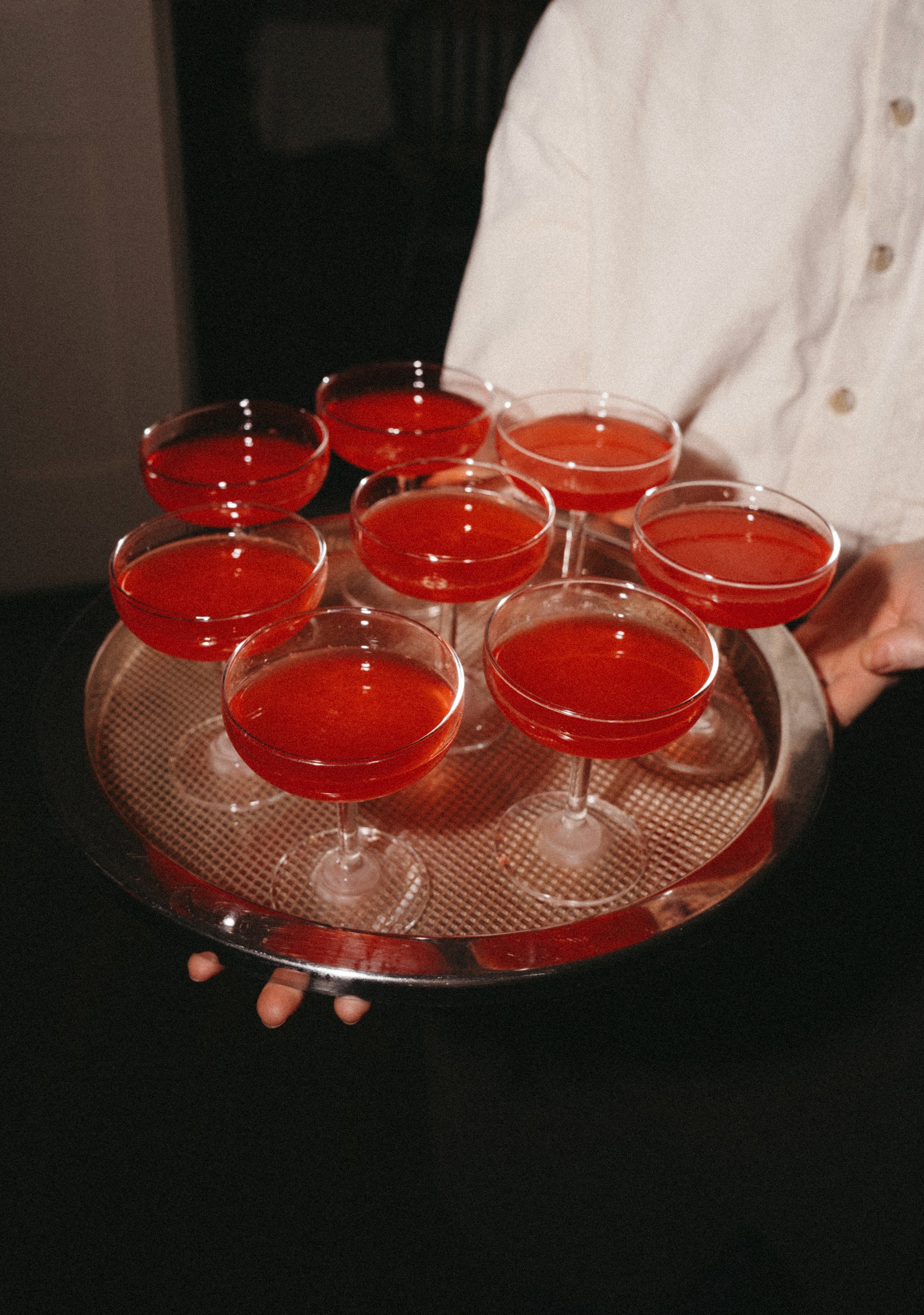 A person holding a silver tray with several red cocktails in coupe glasses.