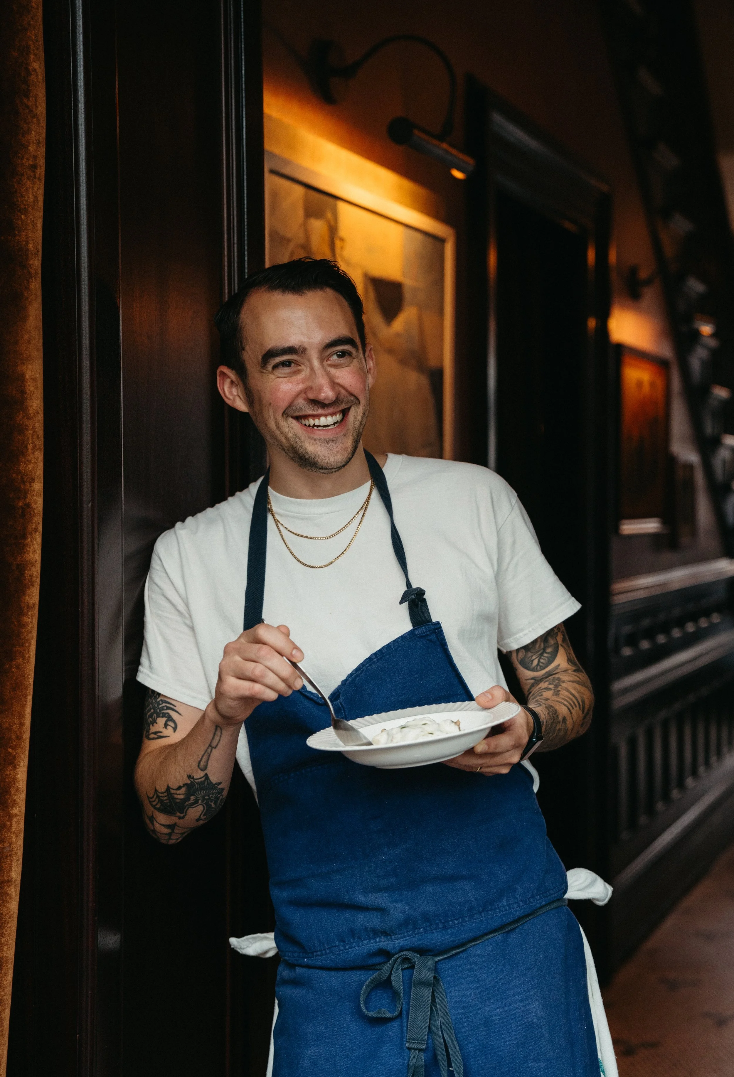 A smiling man wearing a white t-shirt and blue apron, holding a white bowl with food, standing inside a restaurant with dark wood paneling and framed artwork.