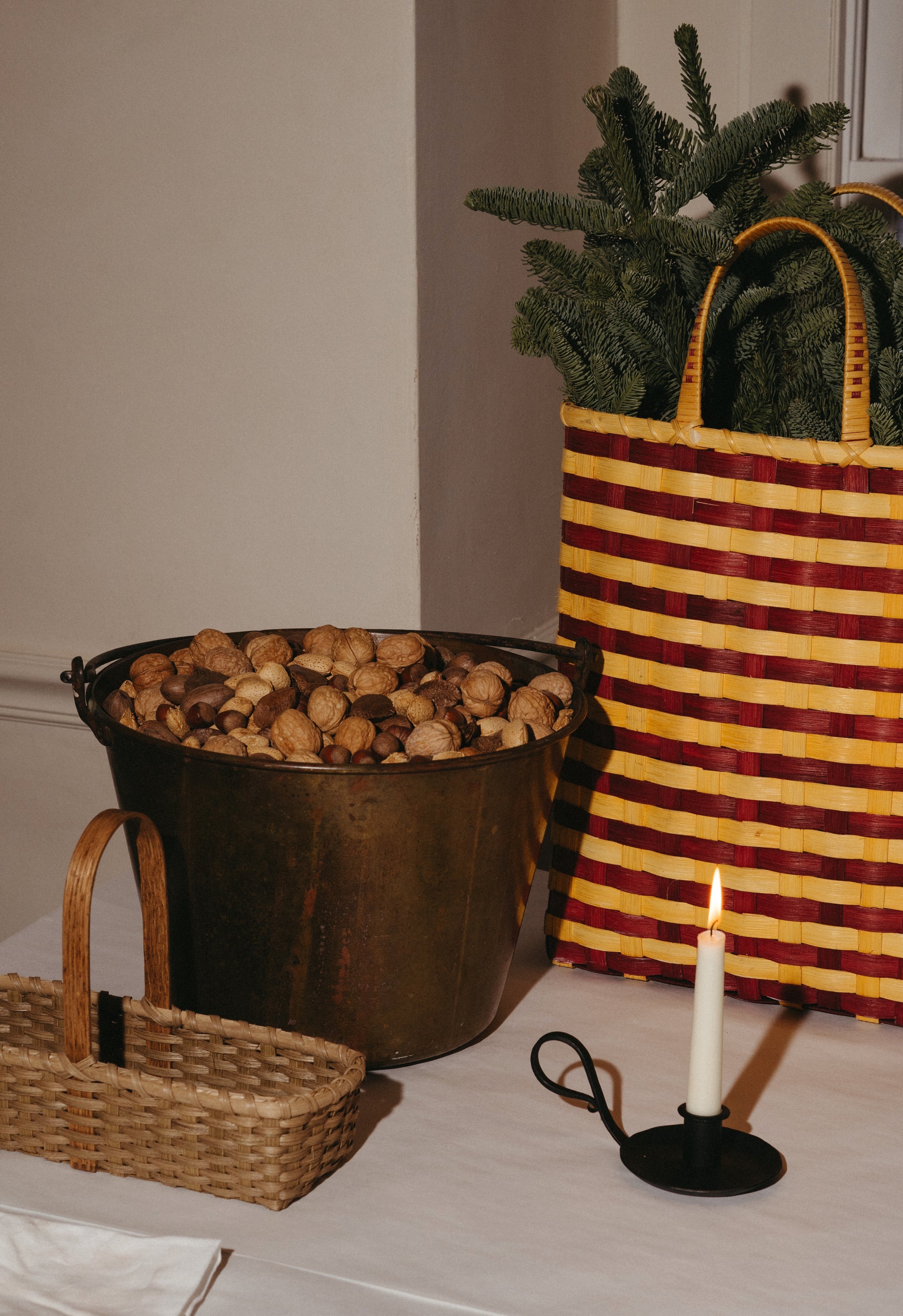 A tabletop display with a basket filled with mixed nuts, a small woven basket, a lit taper candle, a potted pine tree in a red and yellow striped basket, and part of a white tablecloth.