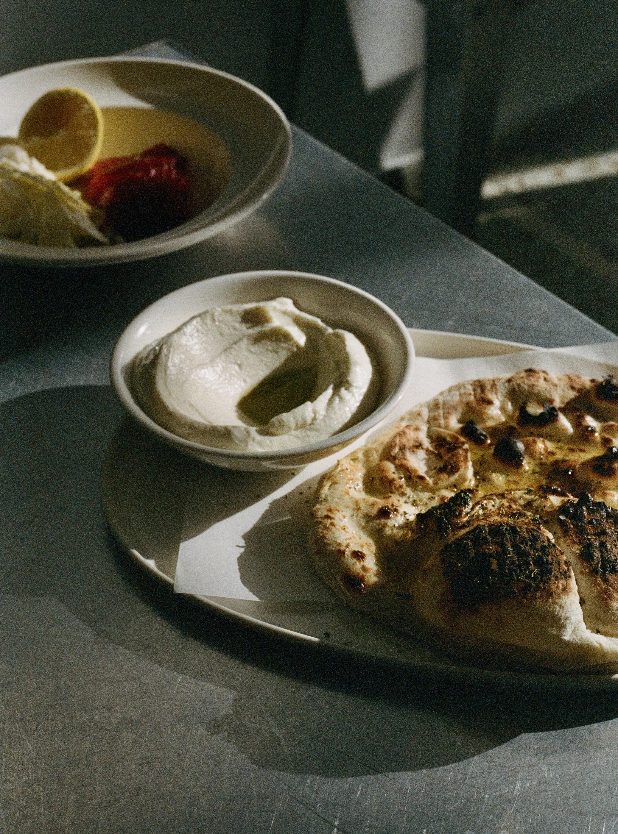 A pita with herbs and olive oil, a bowl of creamy sauce or dip, and a bowl of fish crudo with lemon, on a gray table.