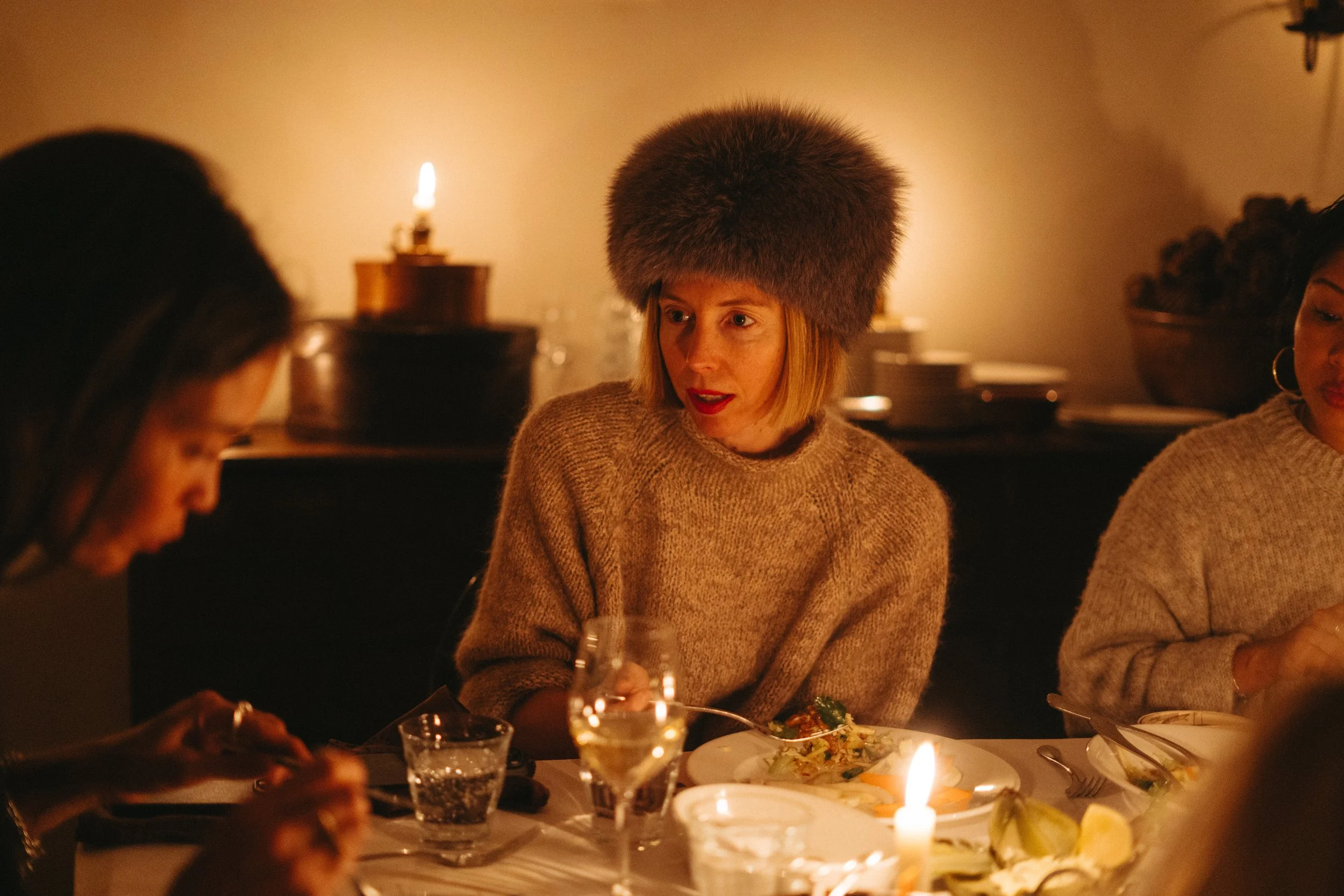 Women dining at a candlelit dinner table in warm lighting, one wearing a large fur hat.
