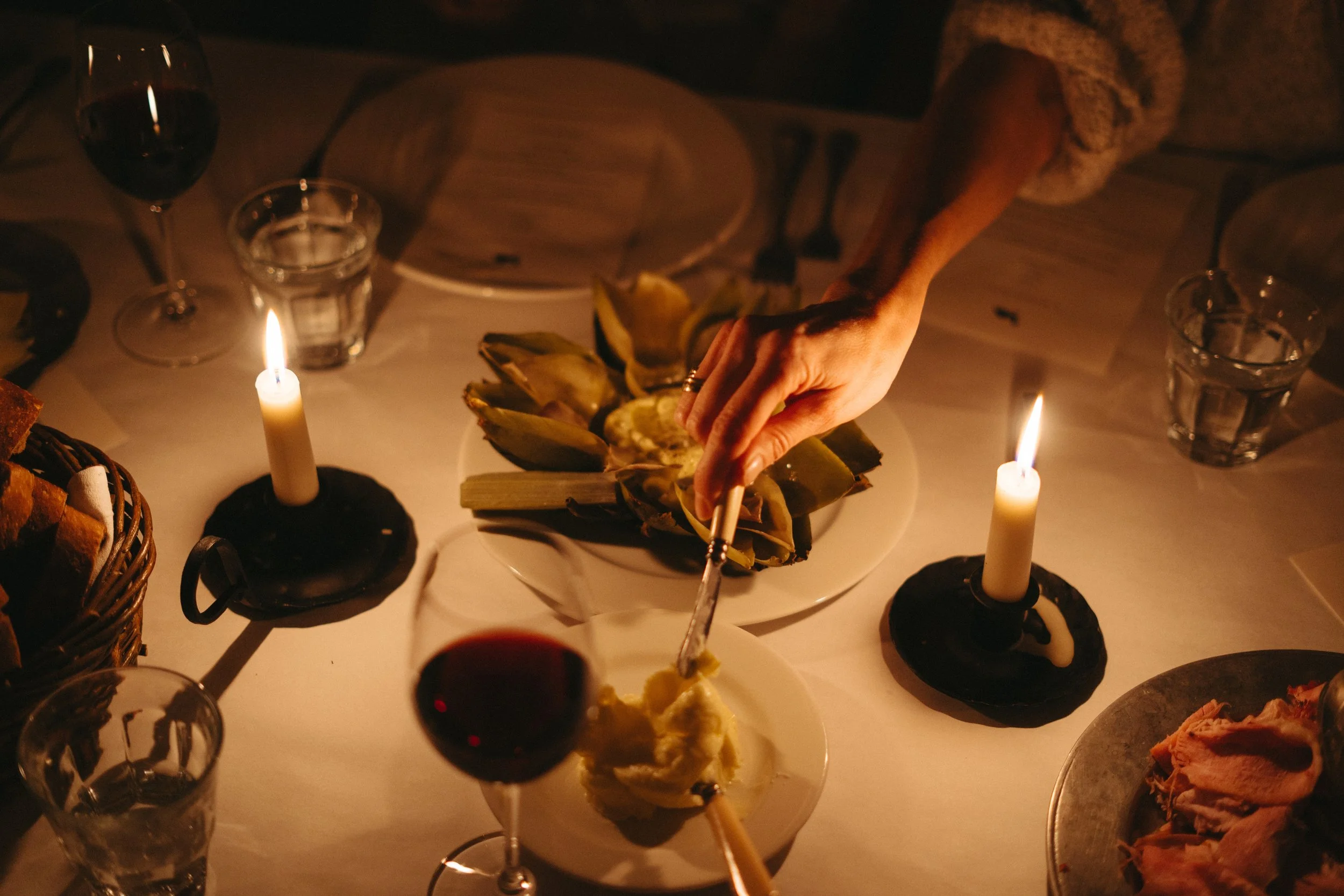 A person serving food at a dinner table illuminated by candles, with plates, glasses of wine and water, and an artichoke on a white plate.