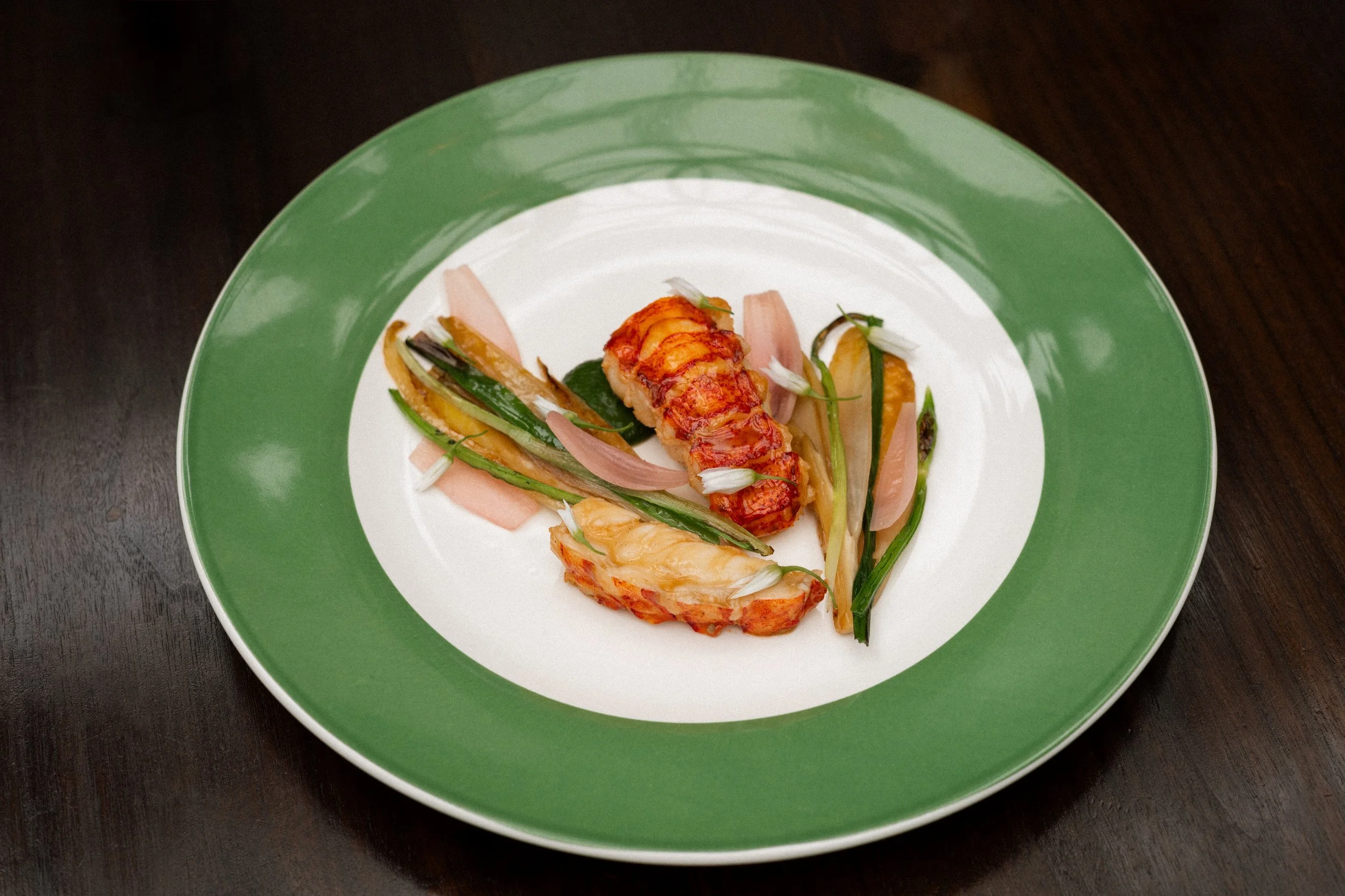 A gourmet plate with lobster tail, grilled vegetables including zucchini, green onions, and onions, garnished with edible flowers, on a green and white plate.