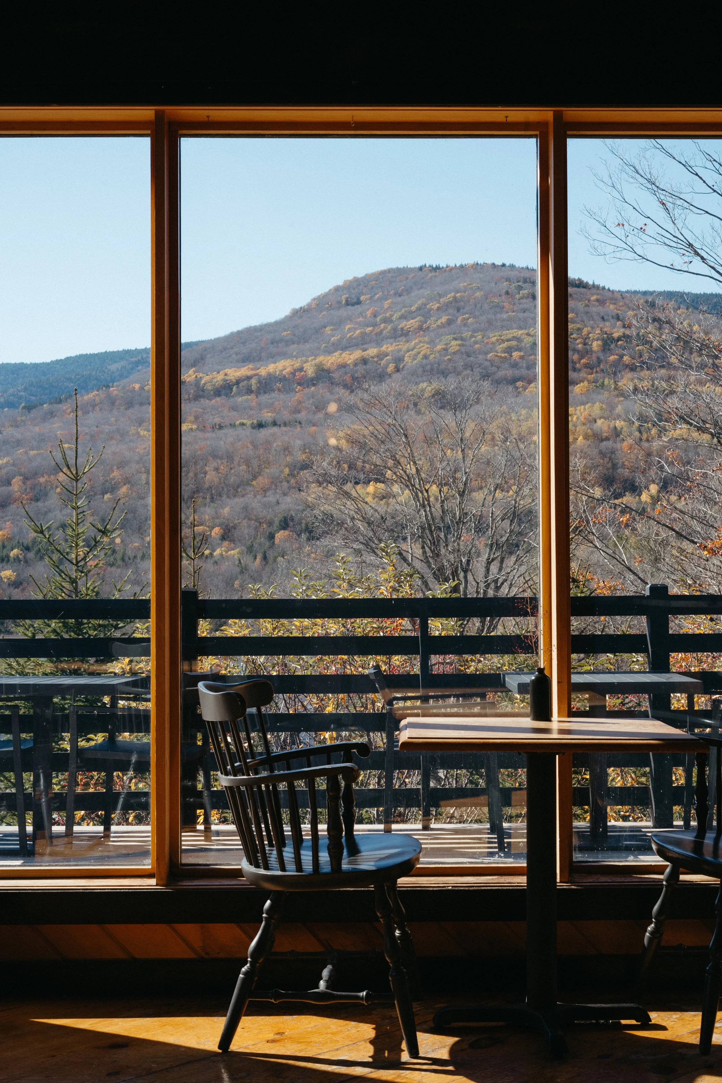 An interior view of a wooden-furnished room with a large window showing a mountain landscape with autumn trees and a clear sky.