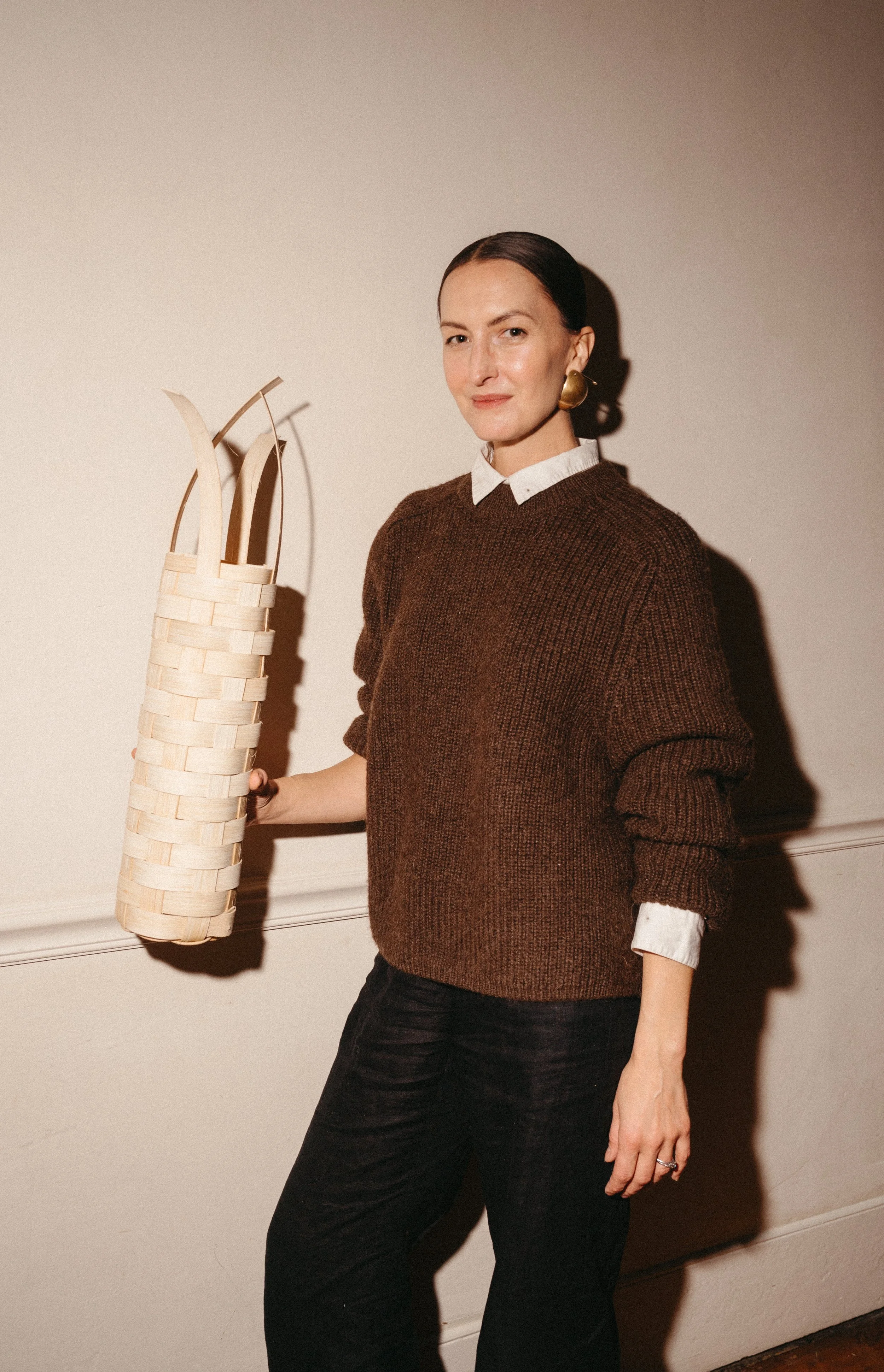 A woman with dark hair pulled back, wearing a white collared shirt under a brown sweater, black pants, and gold earrings, holding a woven basket with a curved handle, standing against a plain wall.