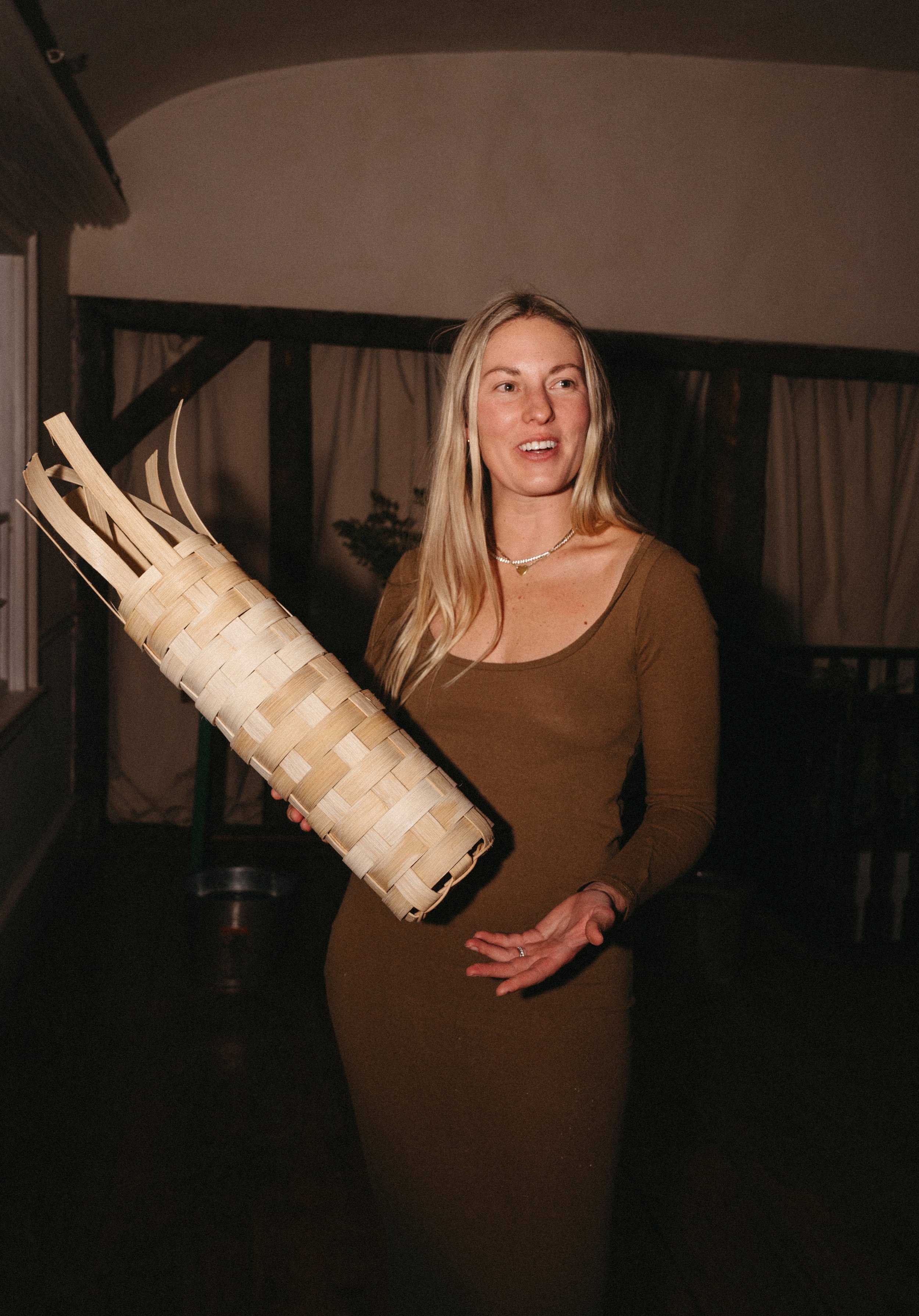A woman with long blonde hair wearing a brown dress, standing indoors, holding a large woven basket.