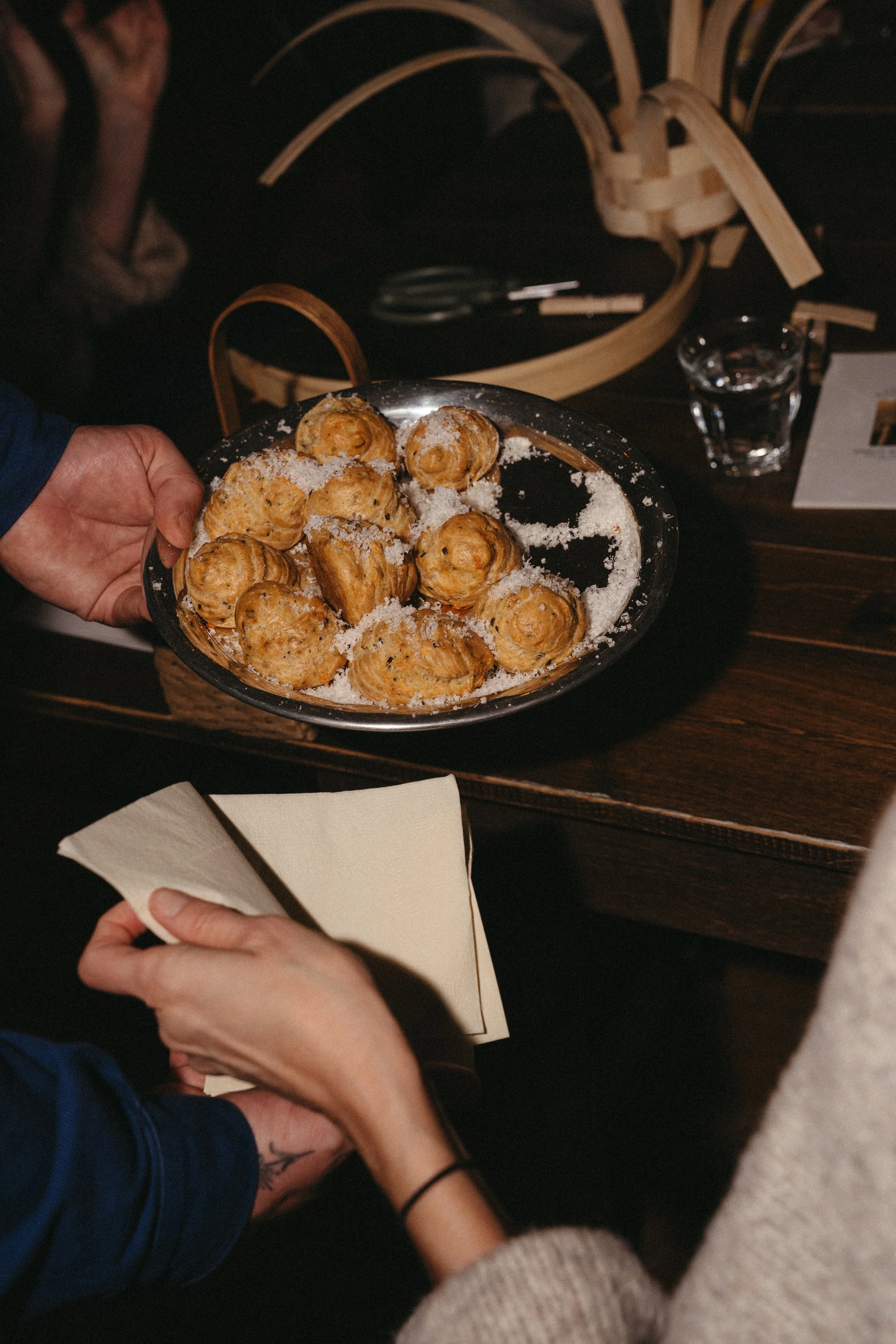 A person holding a plate of baked goods, possibly scones or small bread rolls, dusted with powdered sugar, at a table with napkins, a glass of water, and a basket of wooden utensils in the background.