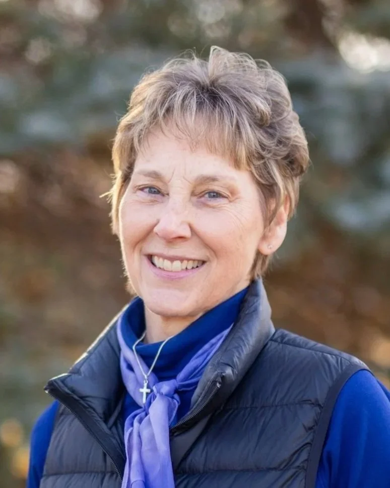 Portrait of a woman smiling outdoors, short light brown hair, wearing a black vest over blue clothing, with a purple scarf and a silver cross necklace.
