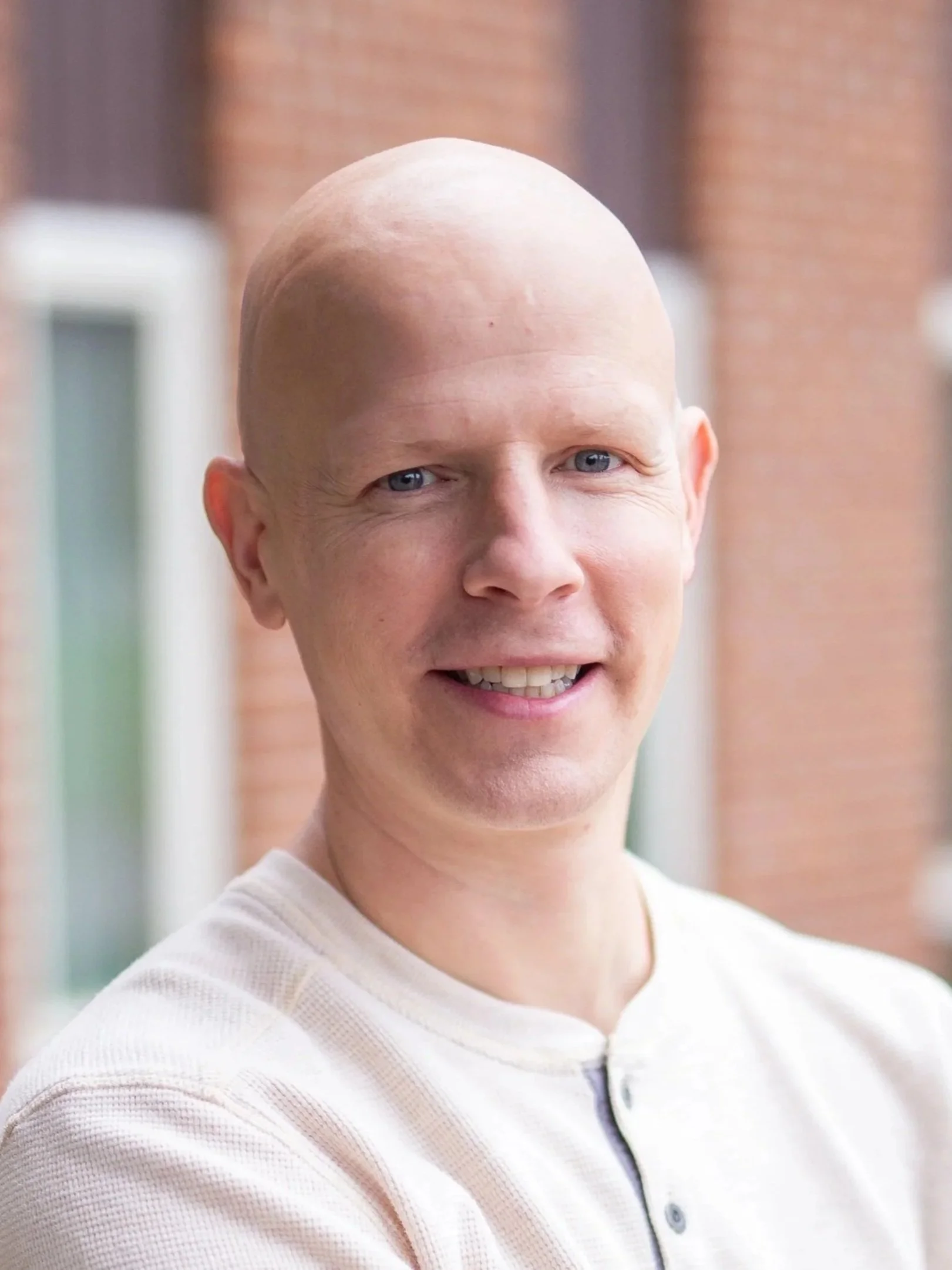 Close-up of a person with a shaved head smiling, wearing a light-colored shirt with a zipper.