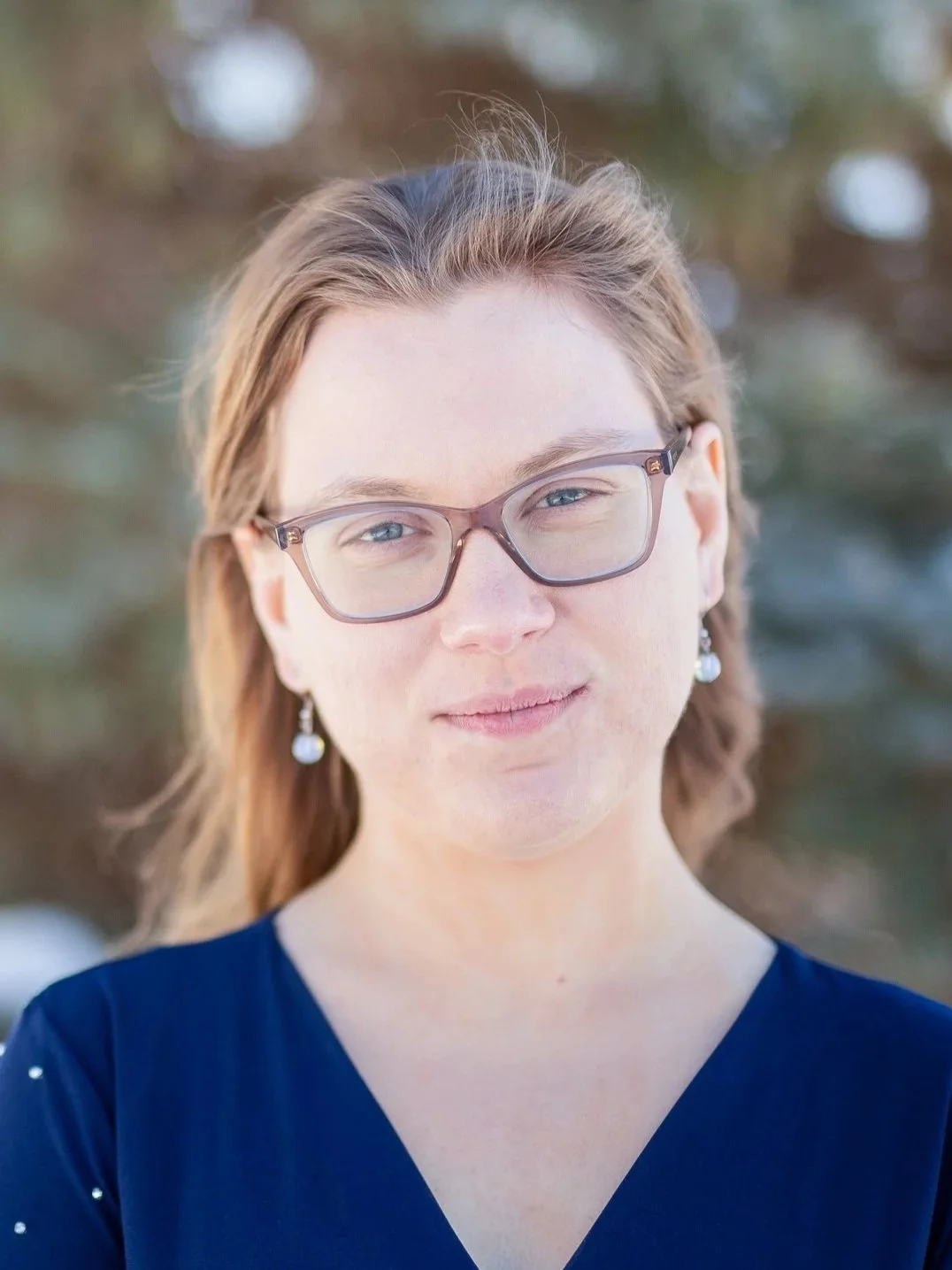 A woman with glasses and pearl earrings, wearing a navy blue top, standing outdoors with blurred trees in the background.