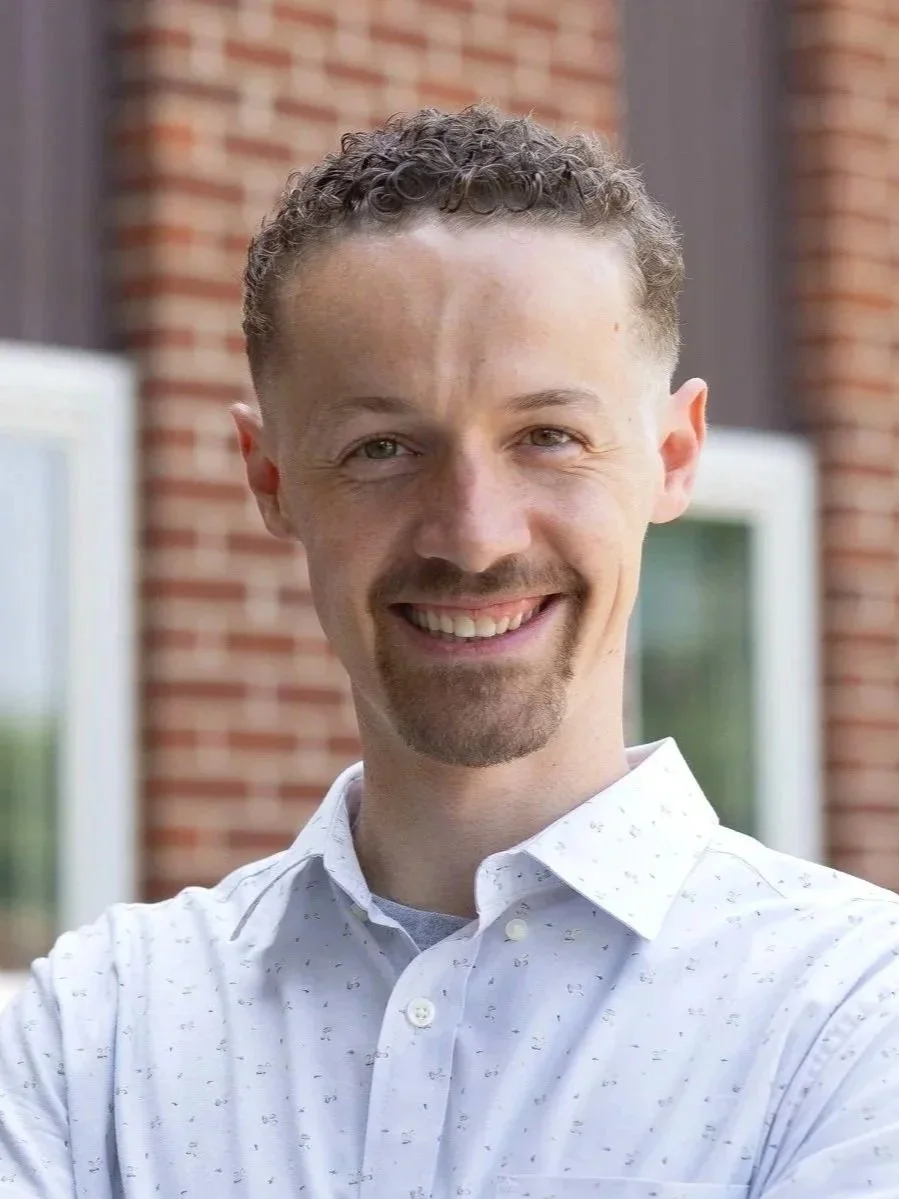 A smiling man with curly hair and a goatee, wearing a white shirt, standing outside in front of a brick building.