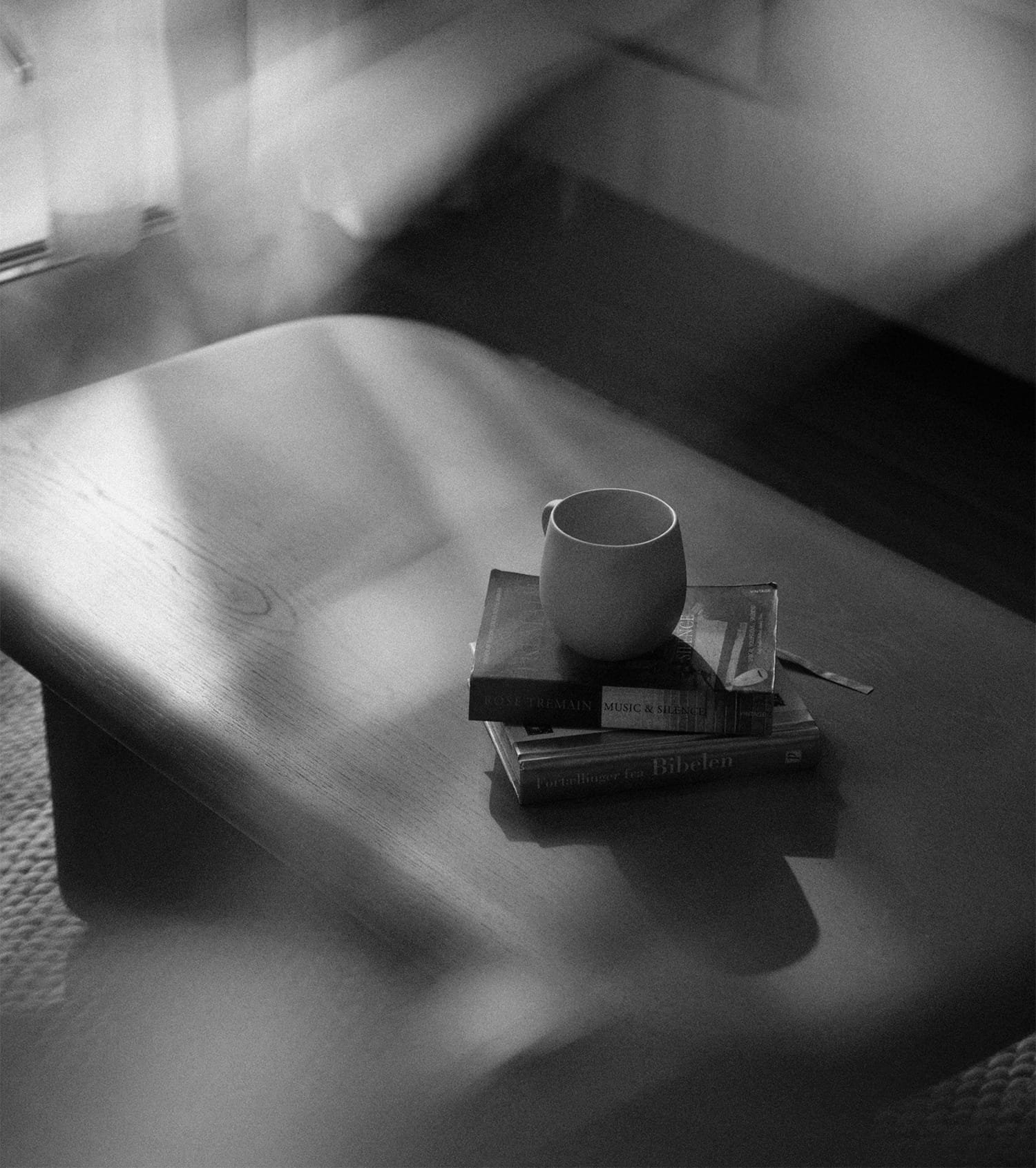 A white mug placed on top of a small stack of books on a wooden table, with a shadow cast across the surface, in black and white.