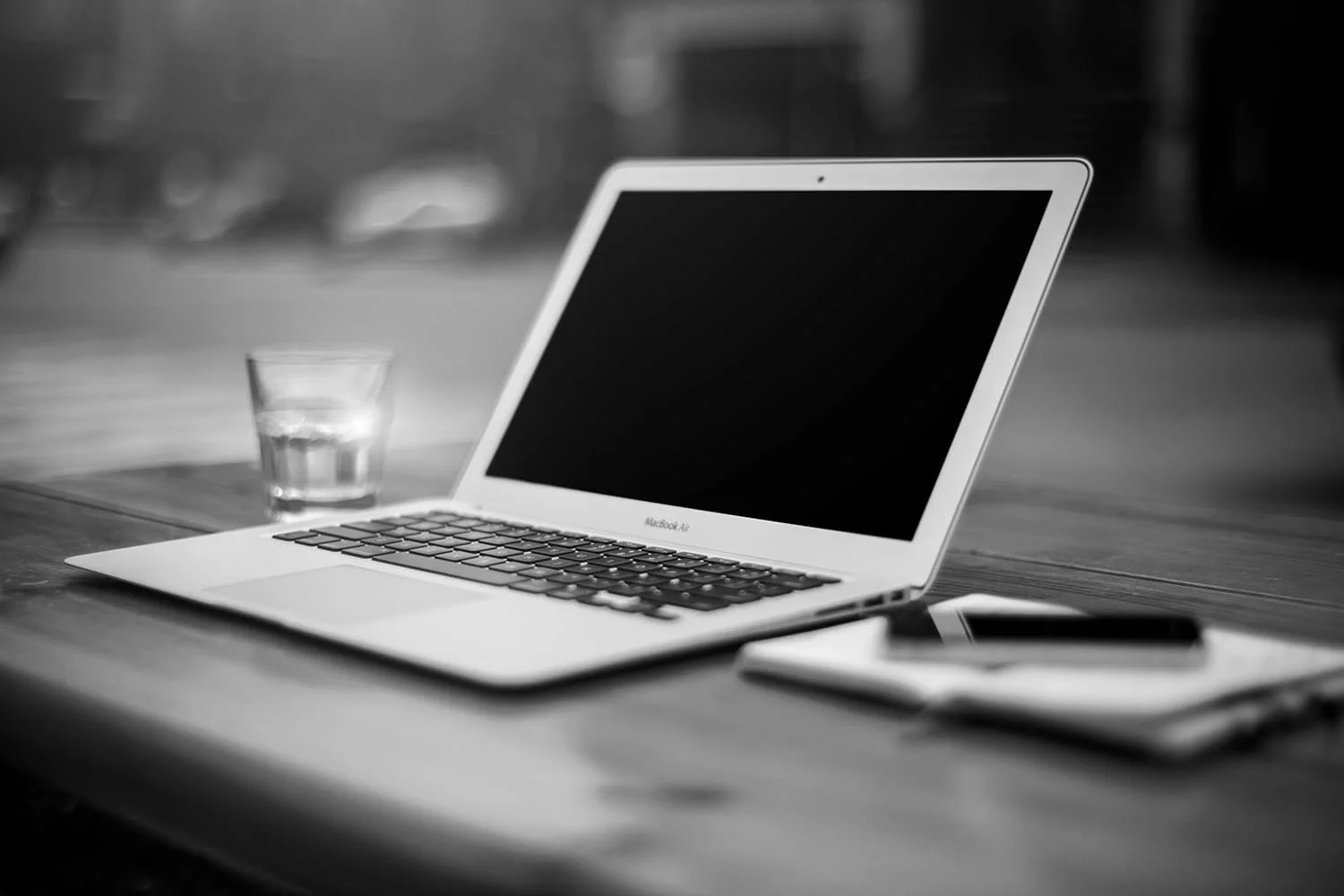Black and white photo of a MacBook Air laptop on a wooden table, with a glass of water, a smartphone, and a notebook nearby.