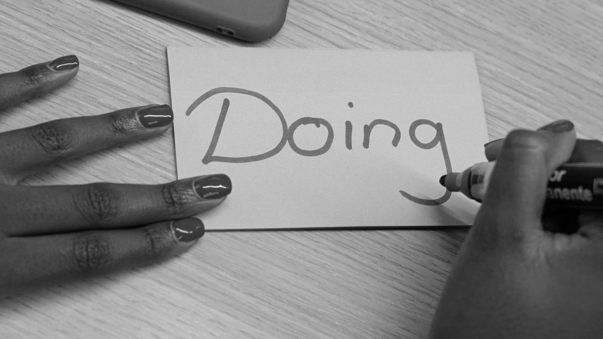 A person with dark painted nails writes the word 'Doing' on a piece of paper with a black marker. The paper is placed on a wooden surface, and a smartphone is partially visible at the top of the image.
