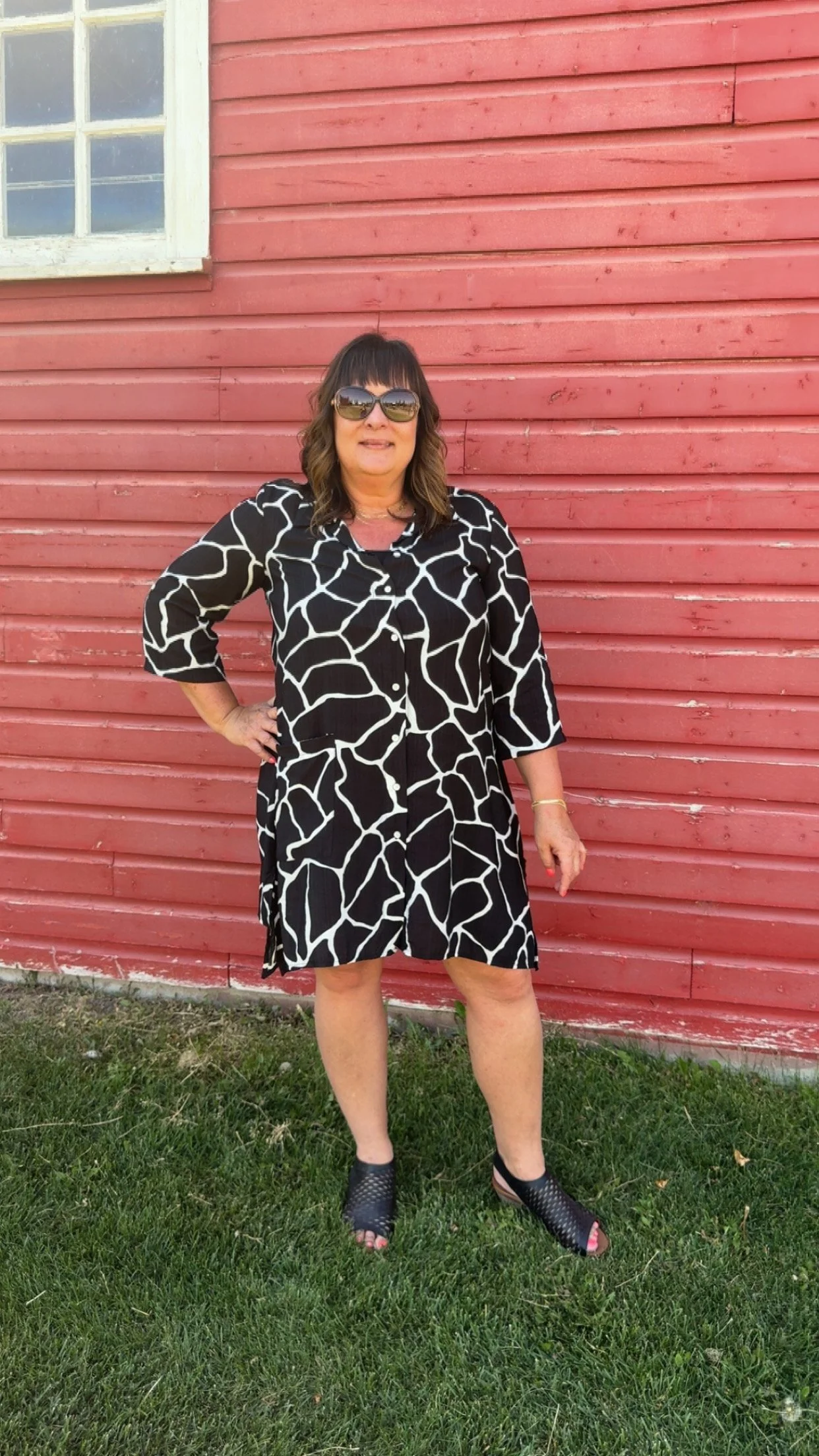 A woman with shoulder-length dark hair wearing sunglasses and a black and white patterned dress, standing outside in front of a red wooden building and green grass.